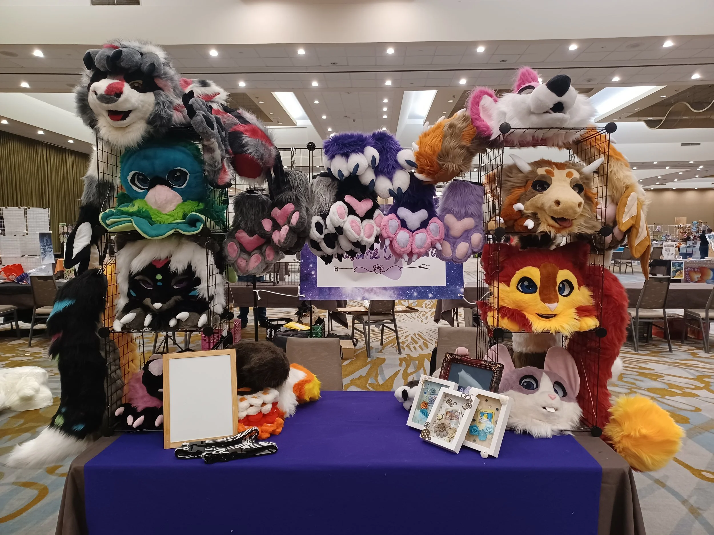 Display table at a convention with colorful, furry animal masks and plush handpaws, including cats, wolves, and other creatures, arranged on a purple cloth and grid walls.