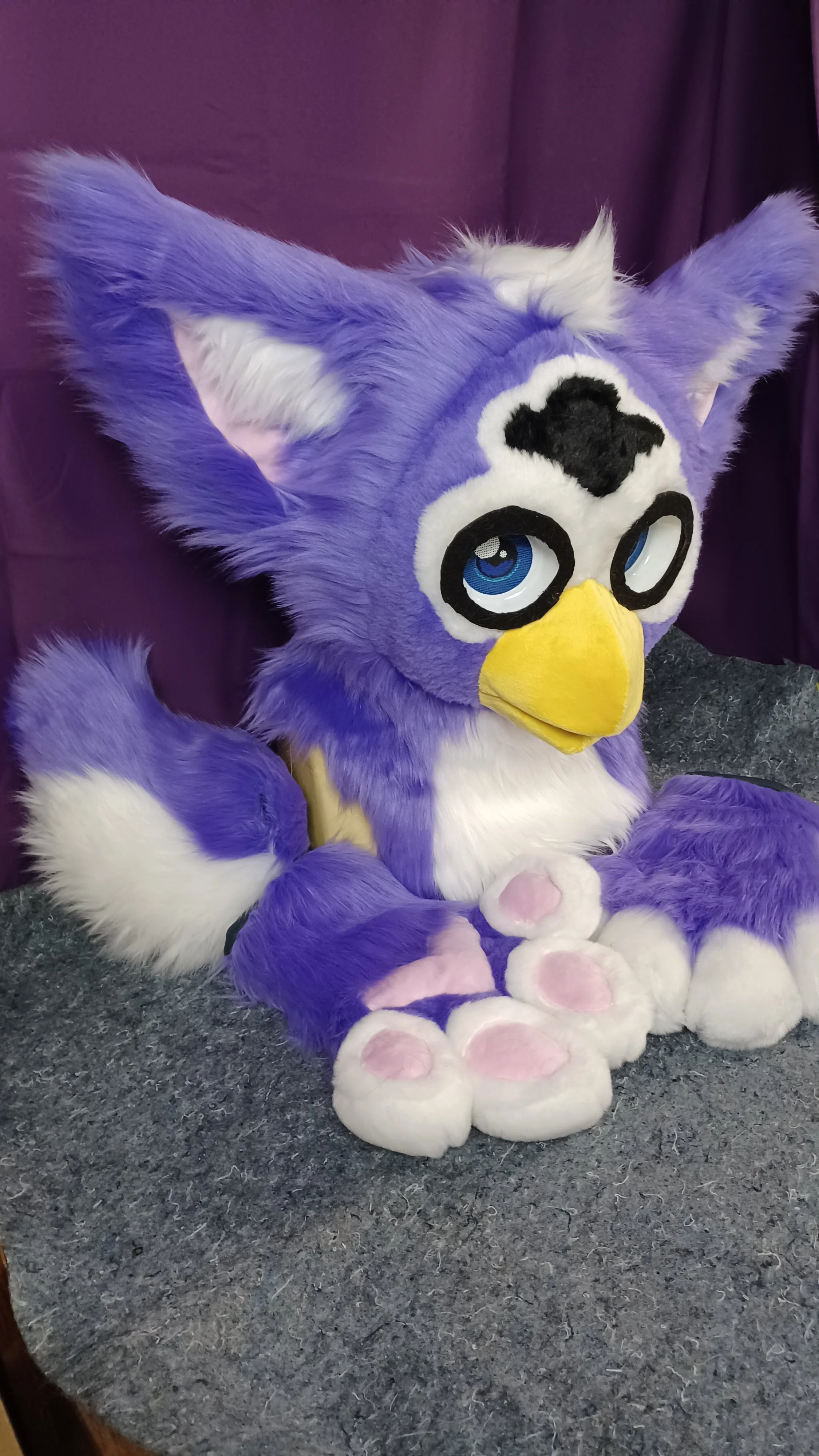 A purple and white furry costume of a cartoon owl character with large blue eyes, yellow beak, and pink paw pads, sitting on a gray textured surface with a purple curtain in the background.
