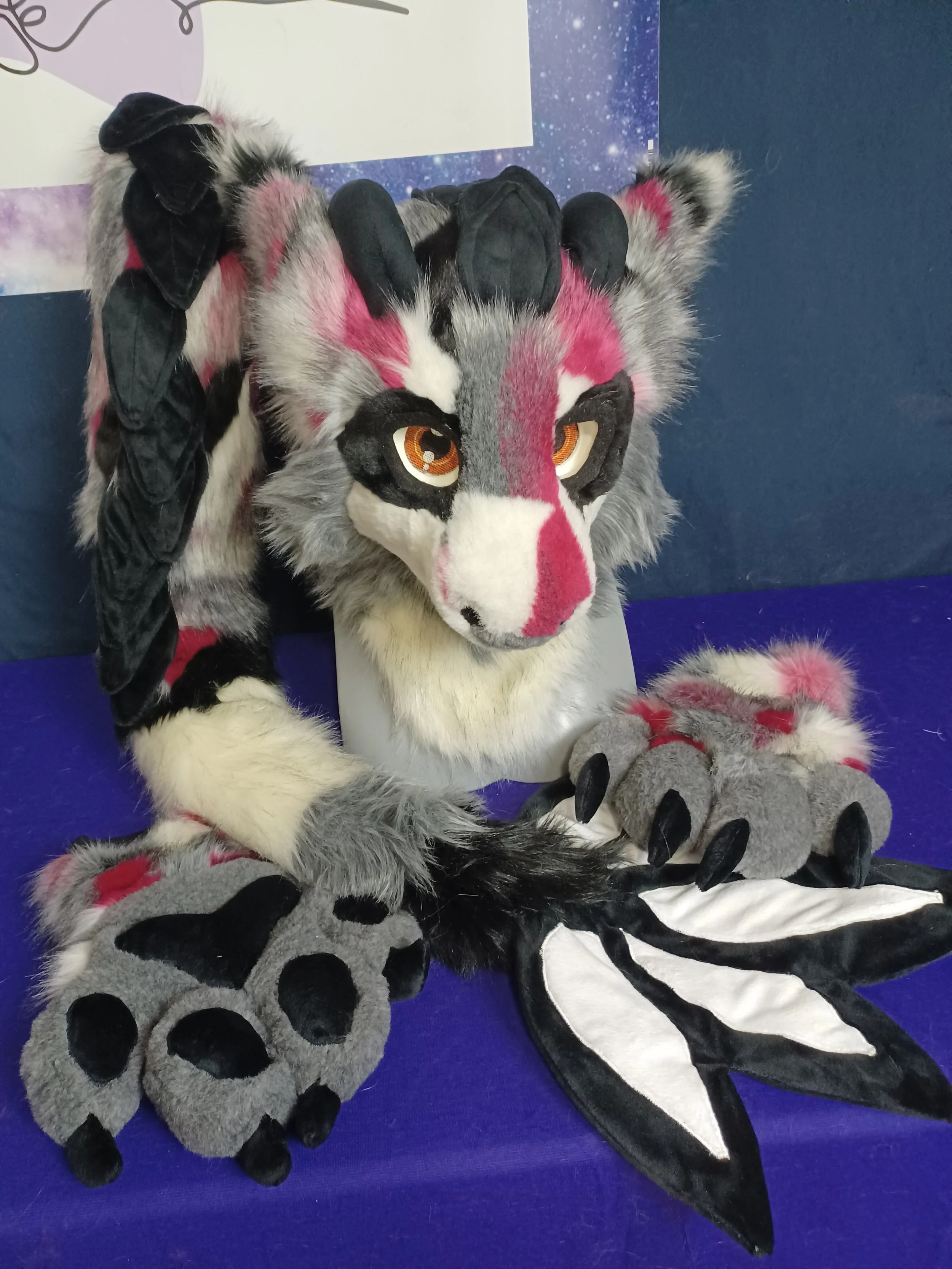 Furry costume of a gray and white wolf with pink and black markings, orange eyes, and a black and white tail; positioned on a blue surface with some plush paw accessories nearby.