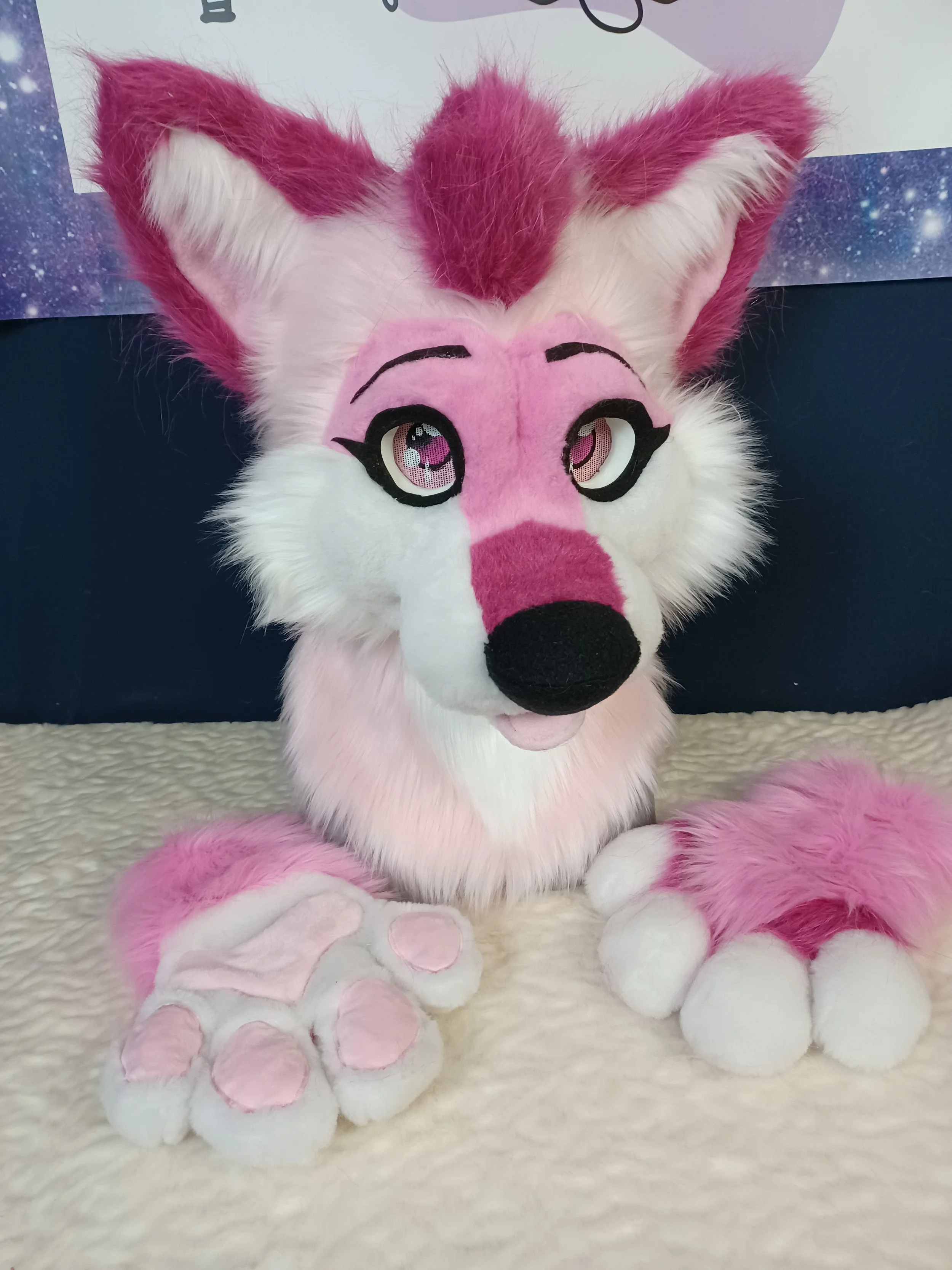 A pink and white furry stuffed animal resembling a fox with large expressive eyes, lying on a soft, cream-colored surface.