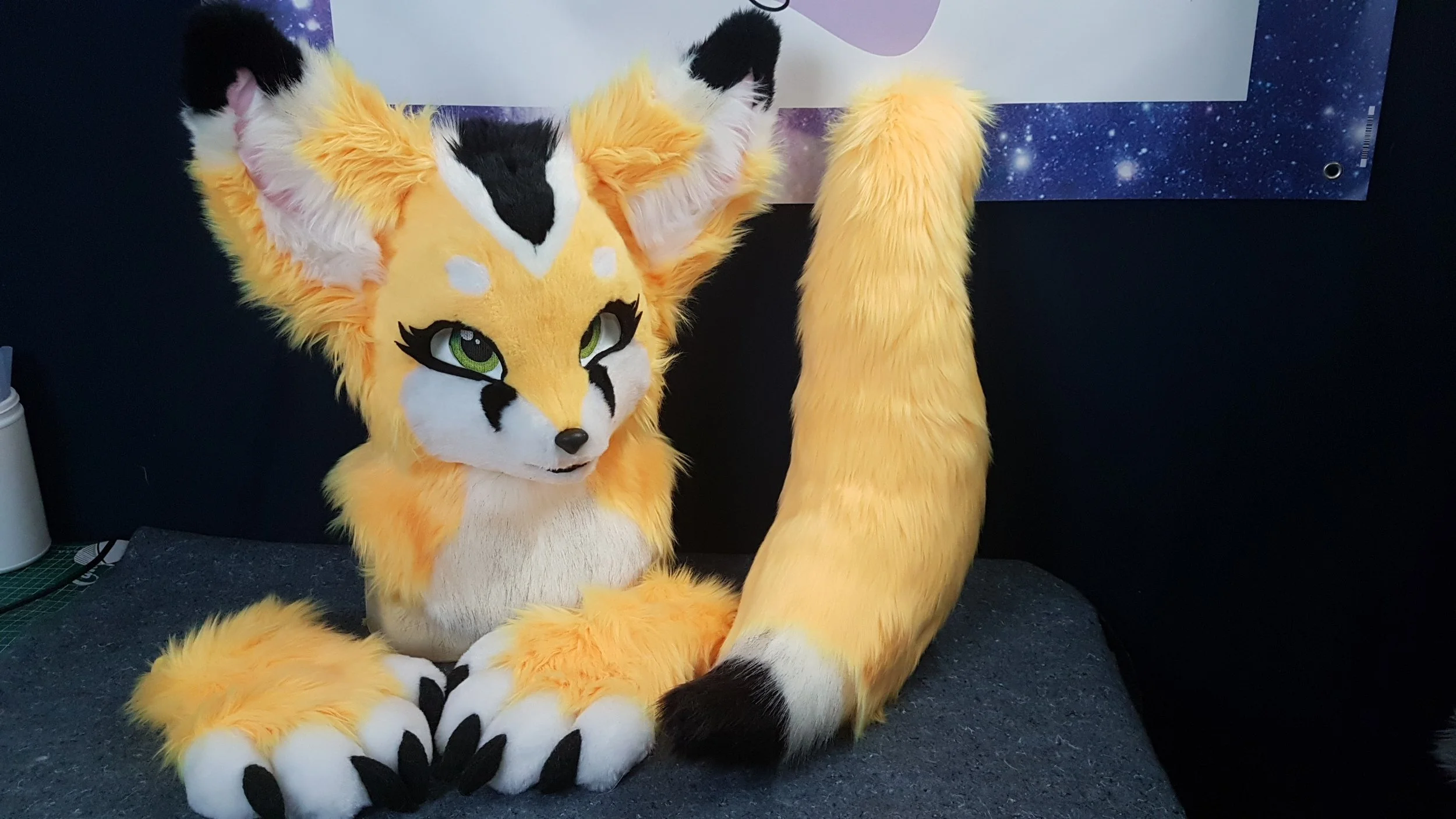 A yellow and white furry fox mascot costume with green eyes, black markings on the face, and large ears, sitting on a dark surface against a blue space-themed background.