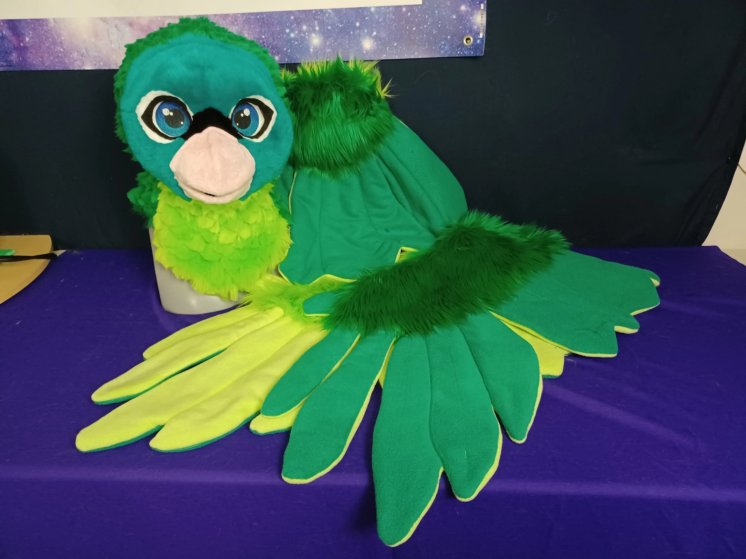 A plush mascot with large blue eyes, a black nose, and green feathers, resembling a bird, displayed on a table with a galaxy-themed banner in the background.
