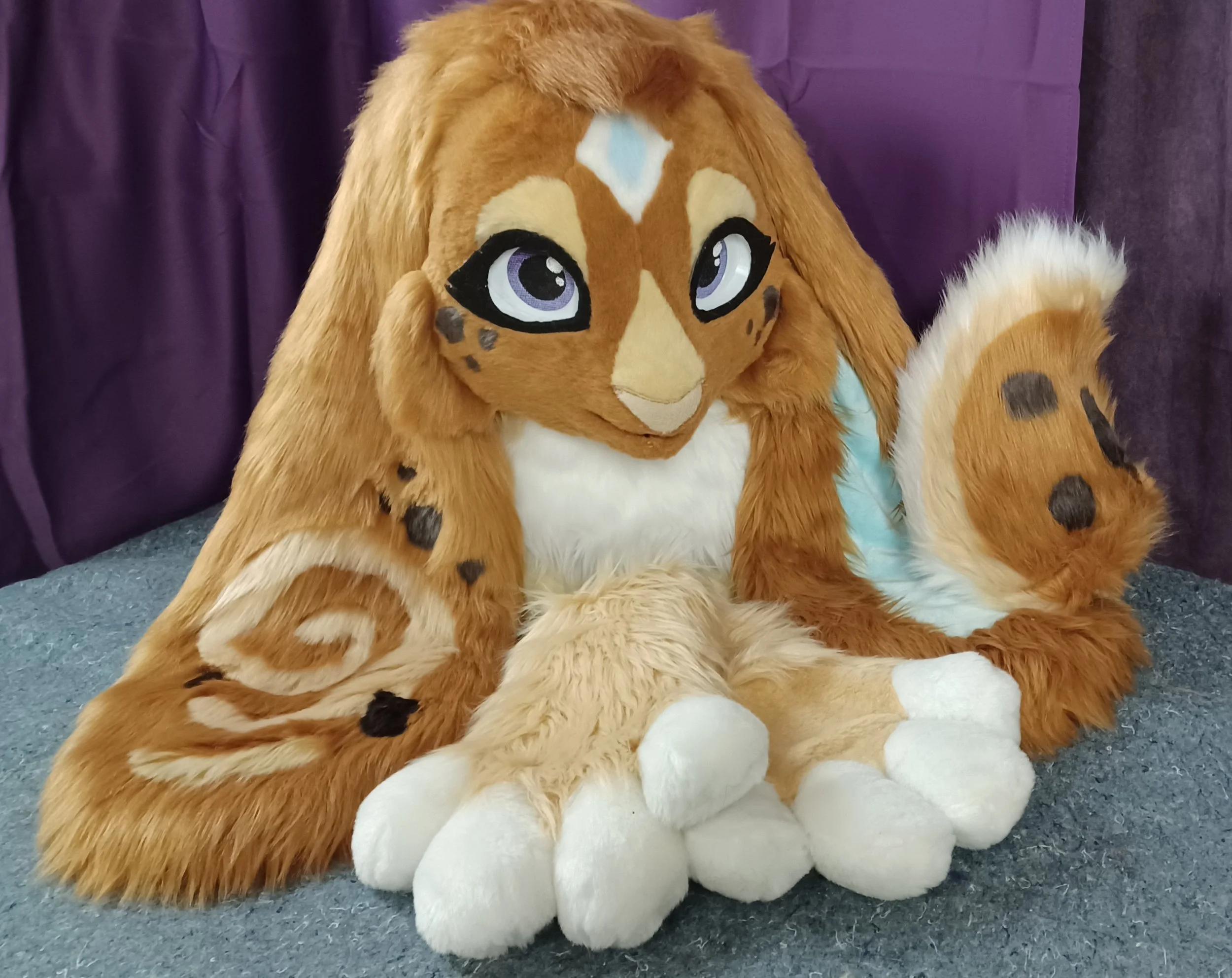 A lop eared bunny fursuit with large blue eyes, a beige face, and a long ears made of golden-brown fur, sitting on a blue carpeted floor with a purple curtain in the background.