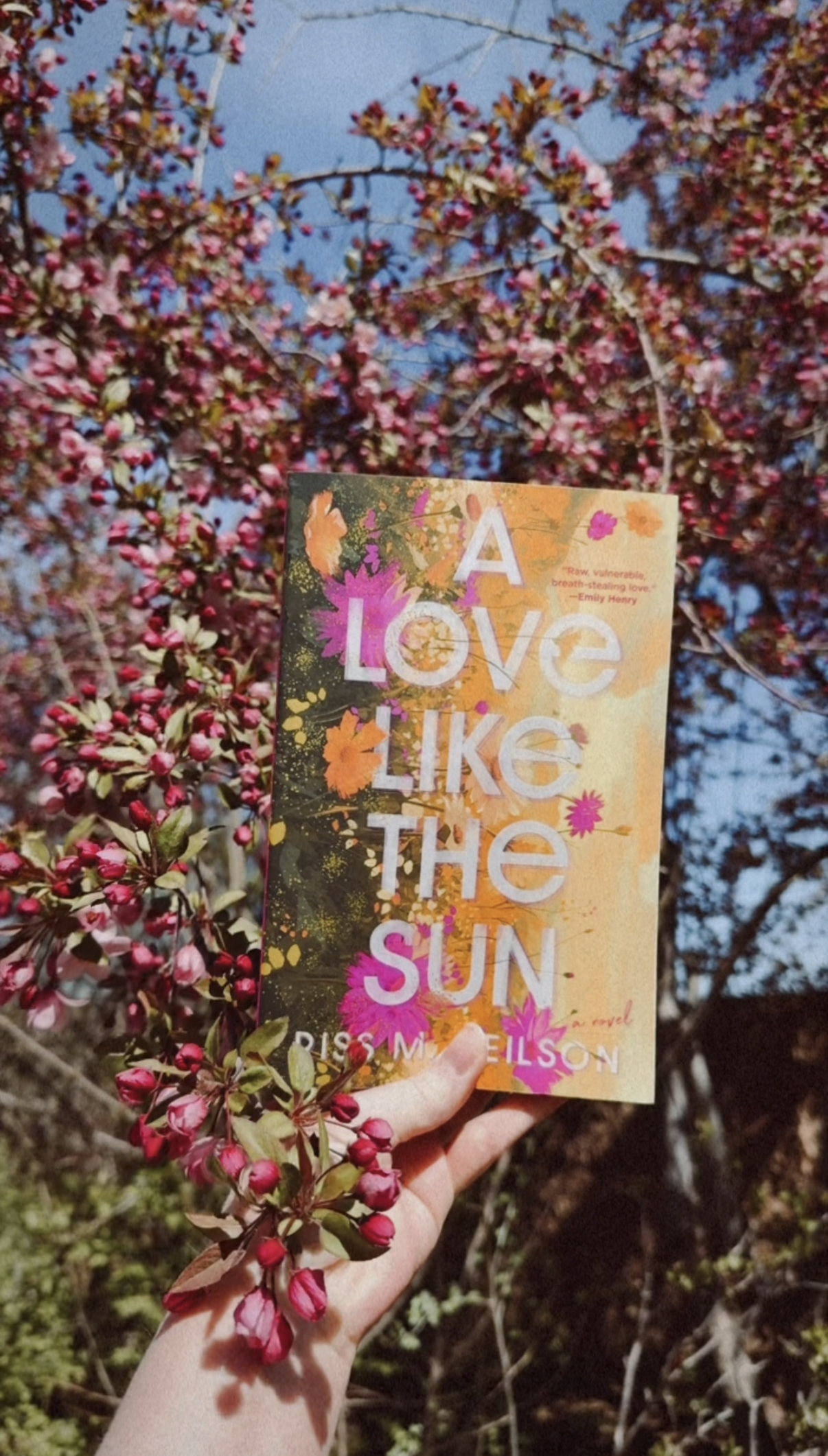 a white hand holding a yellow and pink book up against a bush covered in dark pink flowers blowing in the wind.
