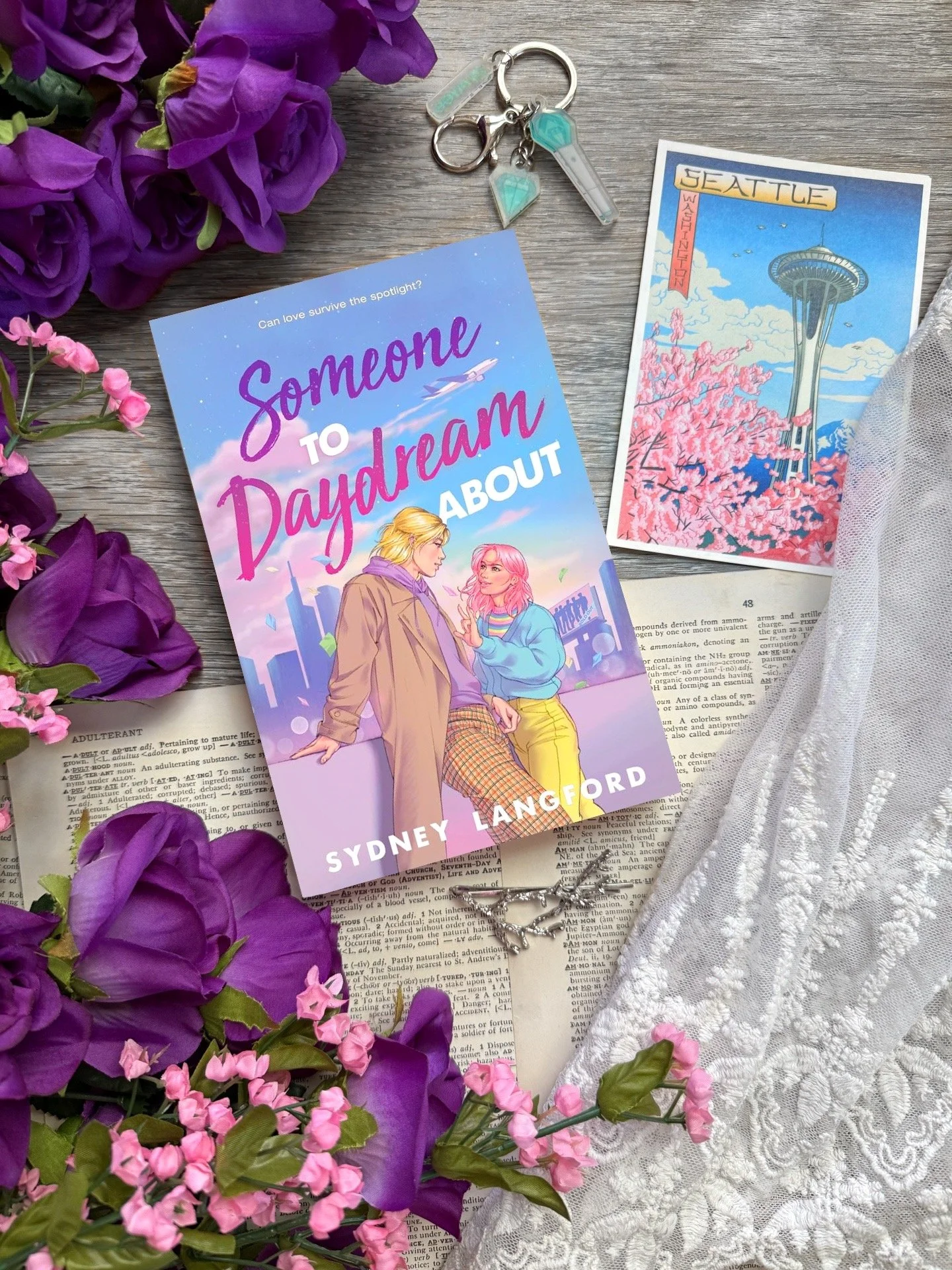 an illustration of a pink haired white teen girl leaning towards a blonde Asian teen and making the ASL sign for “I love you”. A blue city skyline is behind them colored in shades of pink and purple.