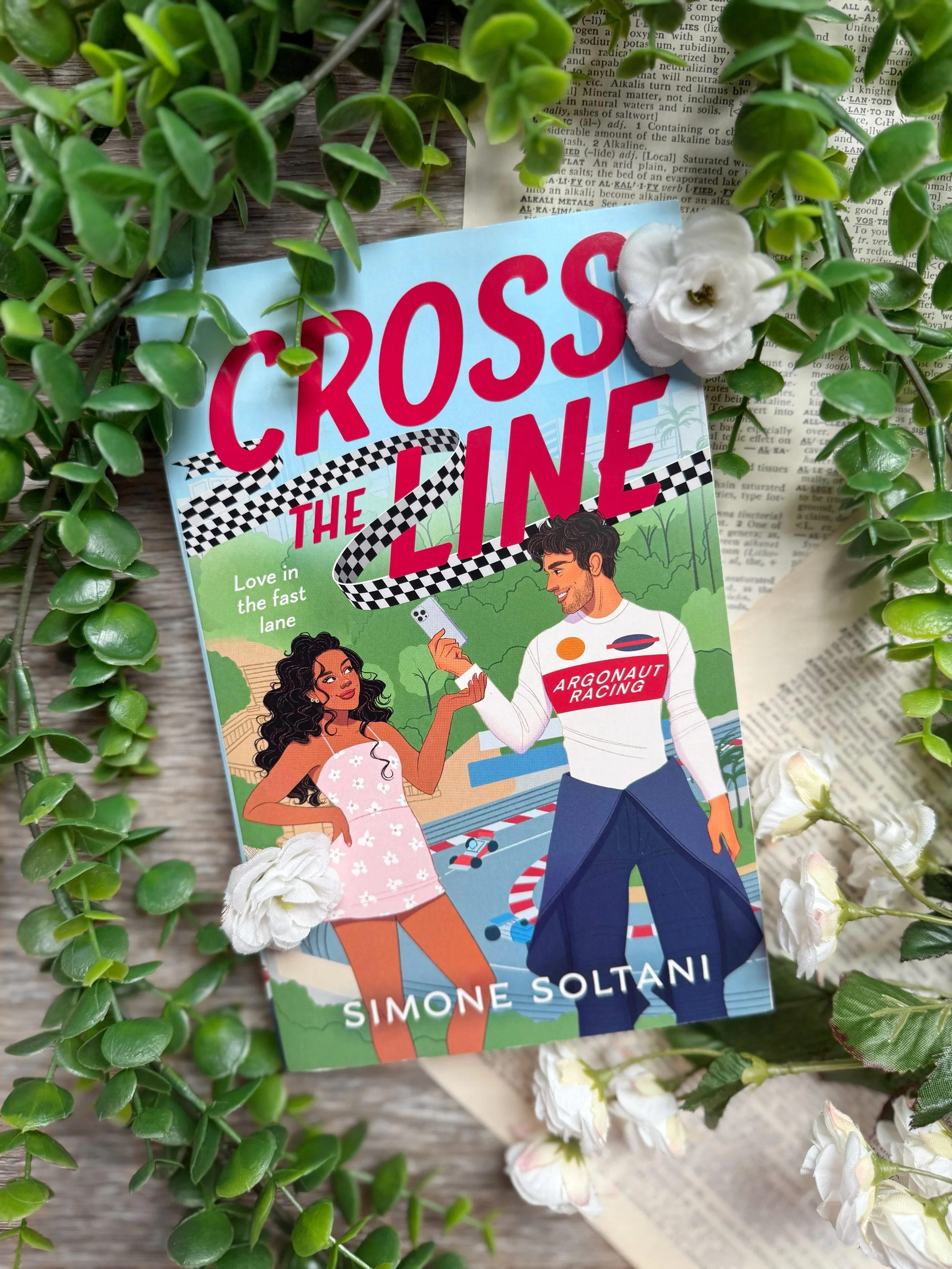 a light blue book cover with an illustration of a Black woman in a pink dress with curly hair gesturing towards a South Asian man in racing gear in front of an F1 racing track.