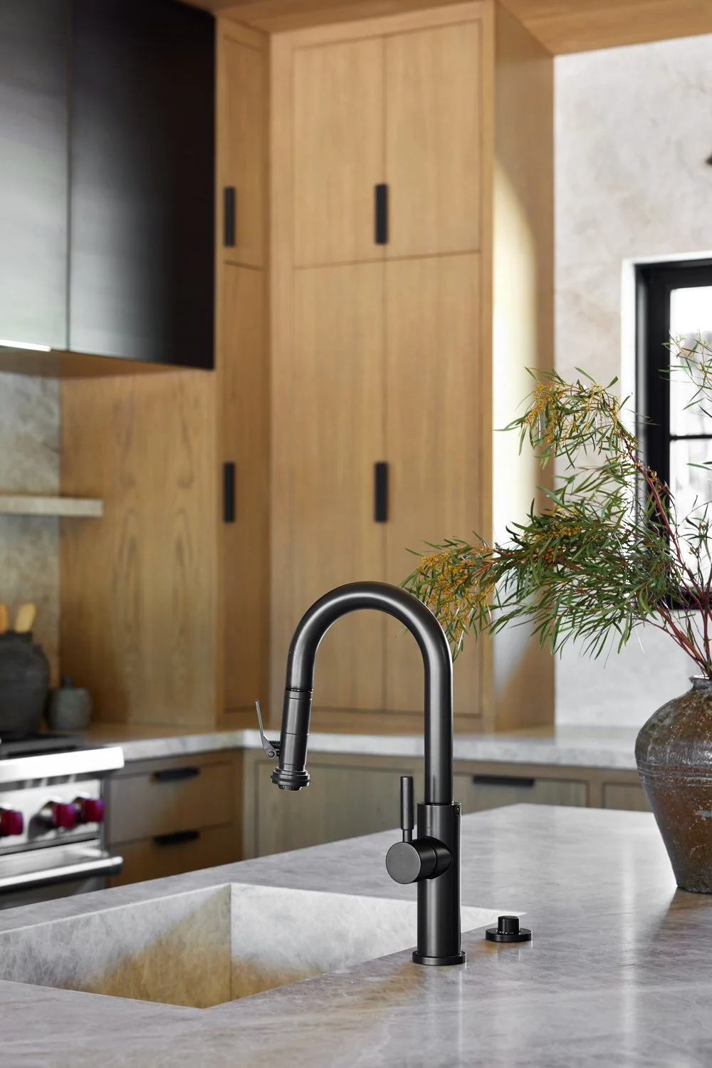 Modern black kitchen faucet over a marble sink in a contemporary kitchen with wooden cabinets and a vase with green foliage.