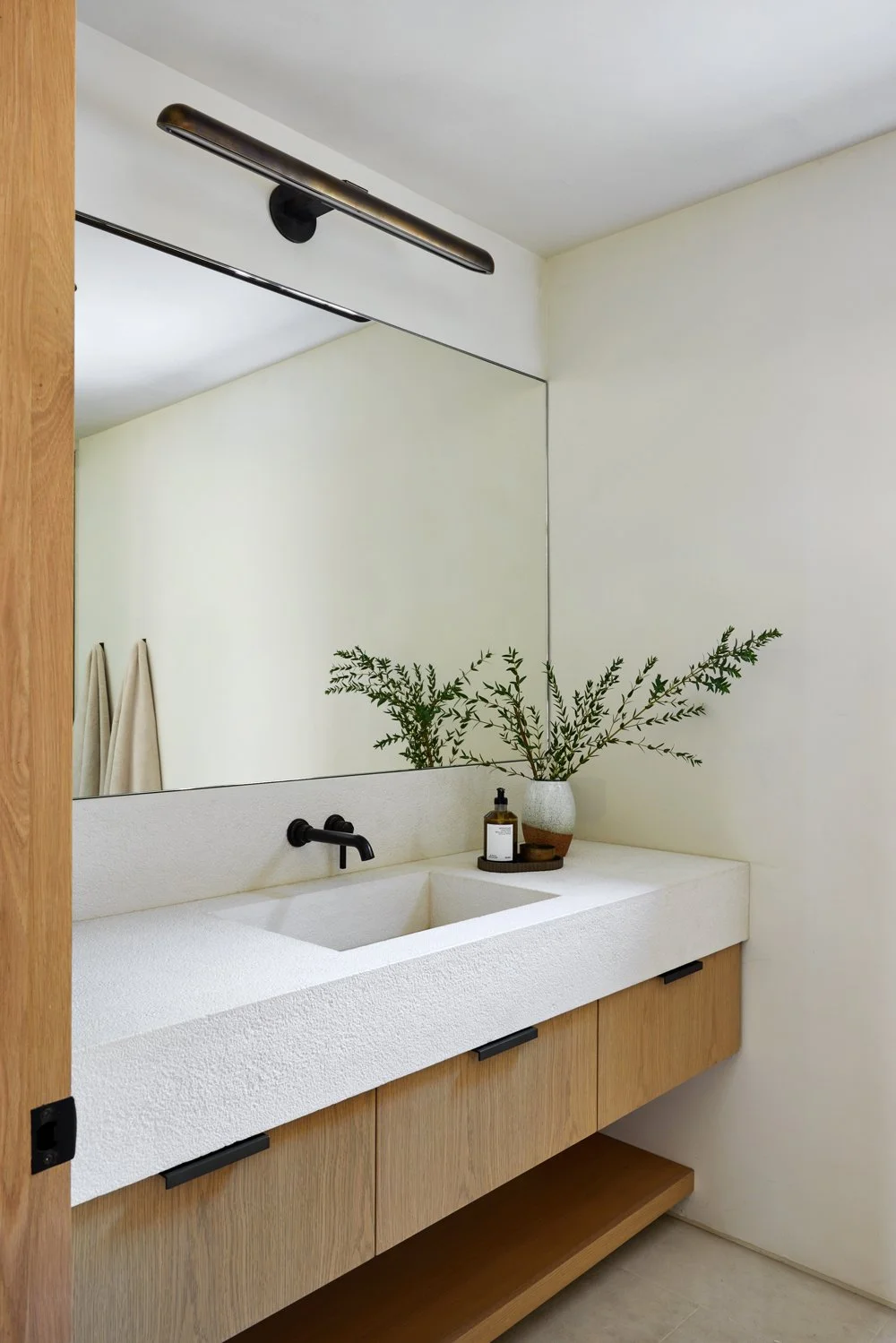 Modern bathroom vanity with a white textured countertop, black faucet, and a large mirror. Decor includes a potted green plant and a small tray with toiletries.