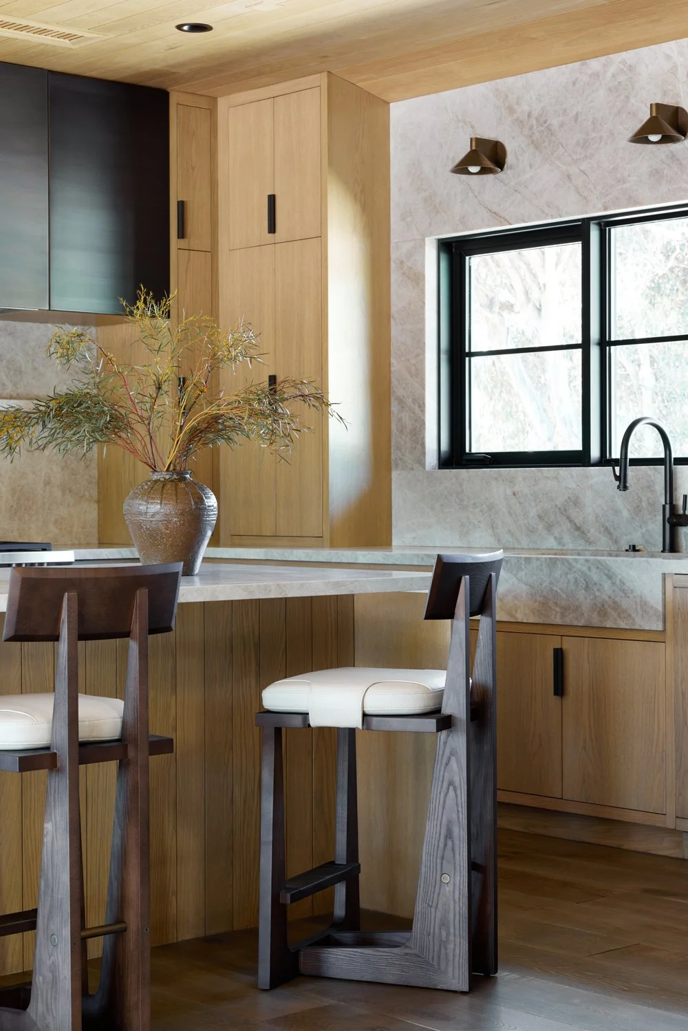 Modern kitchen interior with wooden cabinetry, a marble countertop, black-framed window, and decorative vase with foliage.