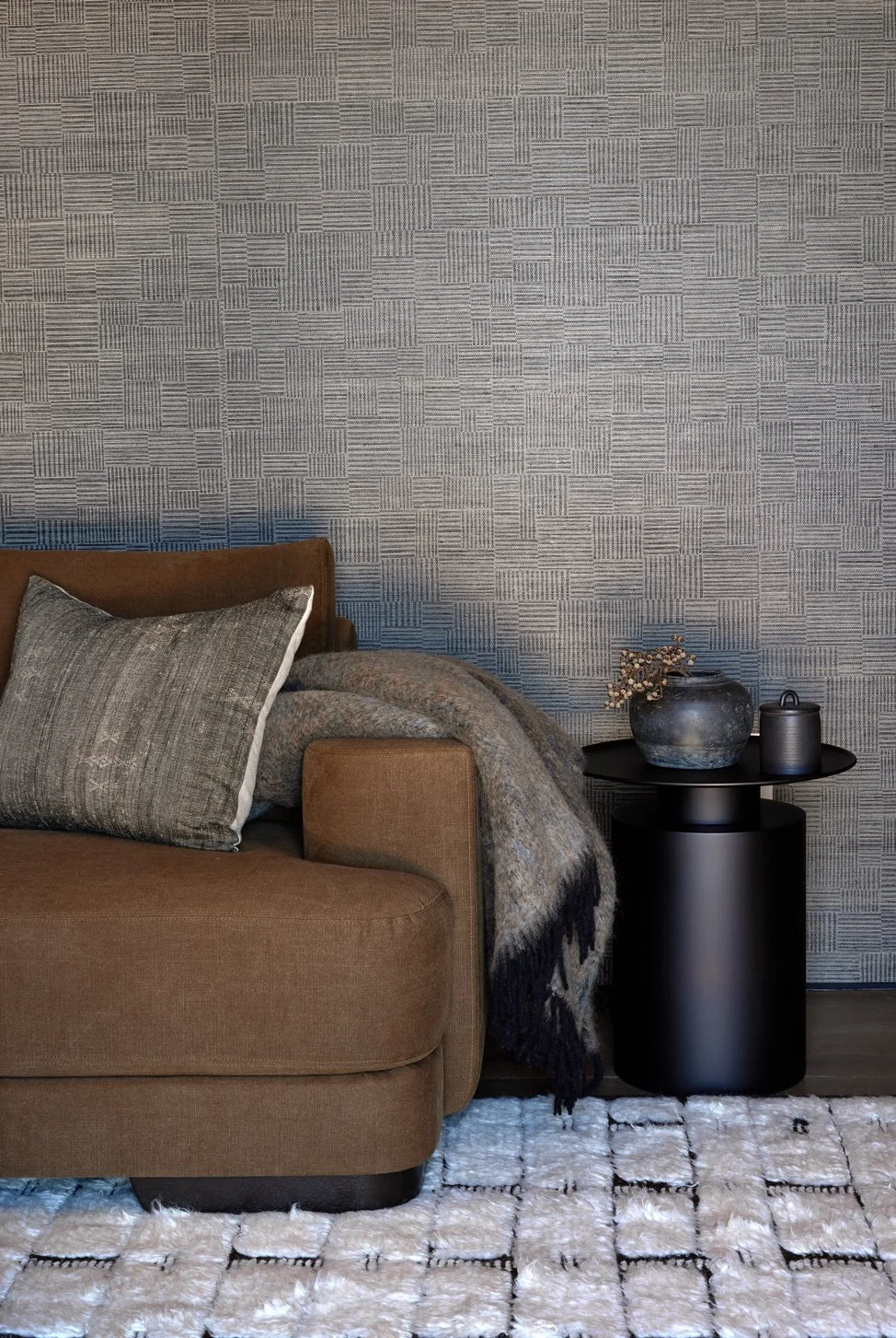 Living room corner with a brown sofa, gray pillow, gray throw blanket, black side table with decorative vases, textured wall, and a white patterned rug.