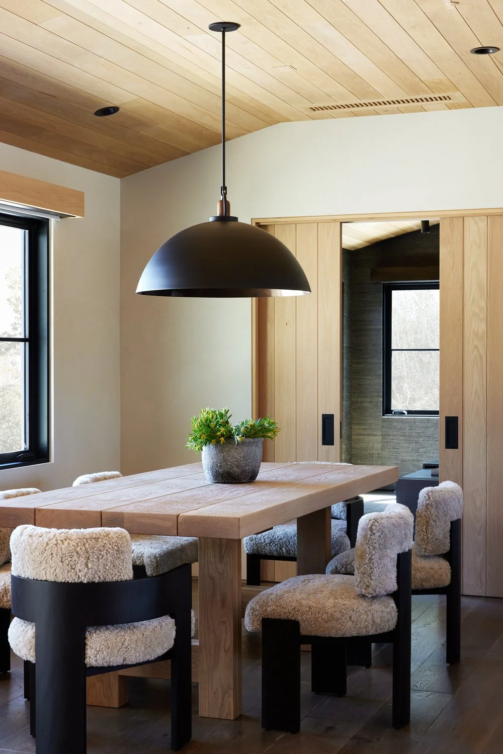A dining room with a wooden table, six chairs with plush sheepskin upholstery, a large black pendant light hanging above the table, and a small green plant in a gray pot as a centerpiece. The room has a wooden ceiling and sliding wooden doors, with w