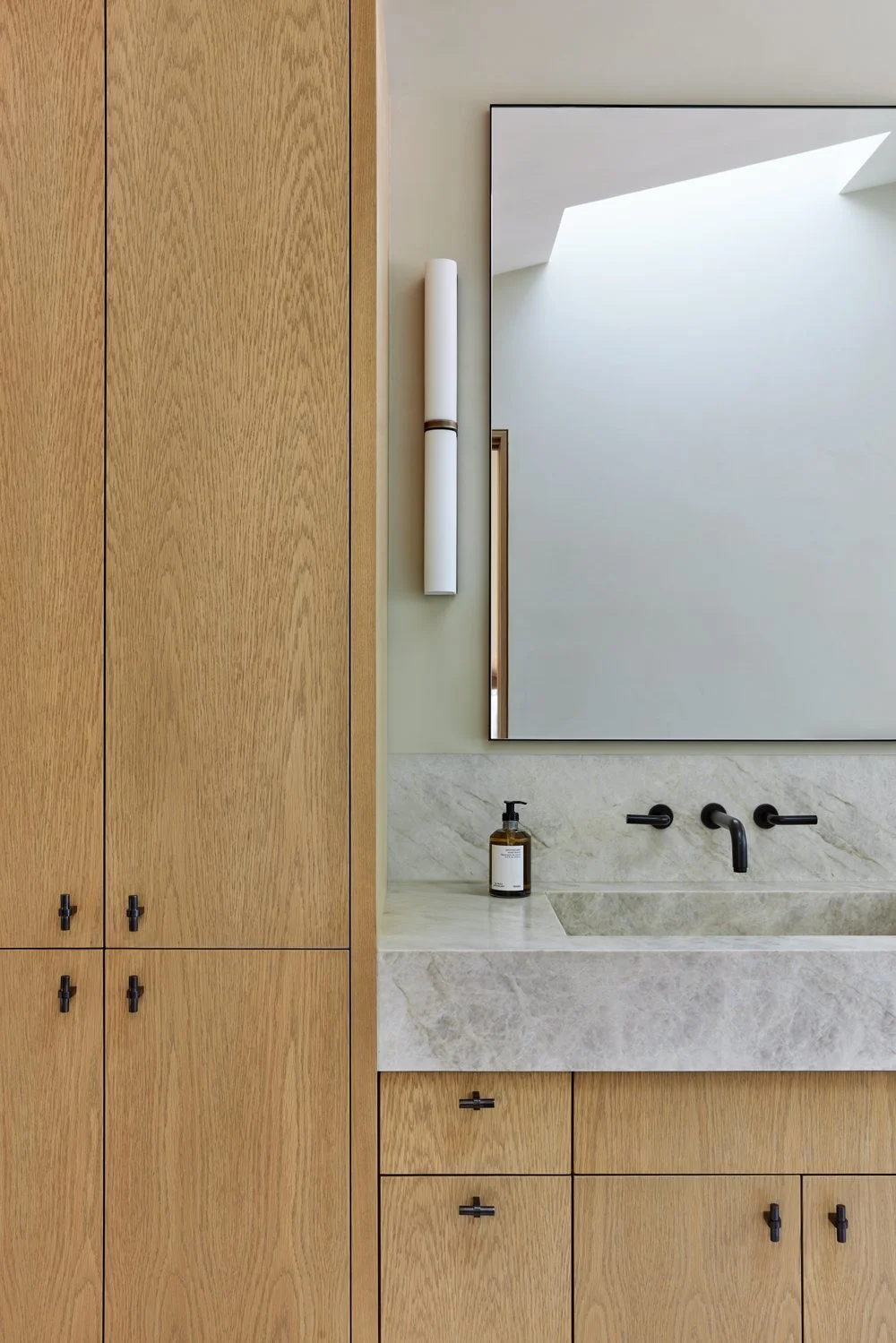 Modern bathroom with wooden cabinets, a marble sink, a large mirror, wall-mounted faucet, soap dispenser, and minimalist lighting.