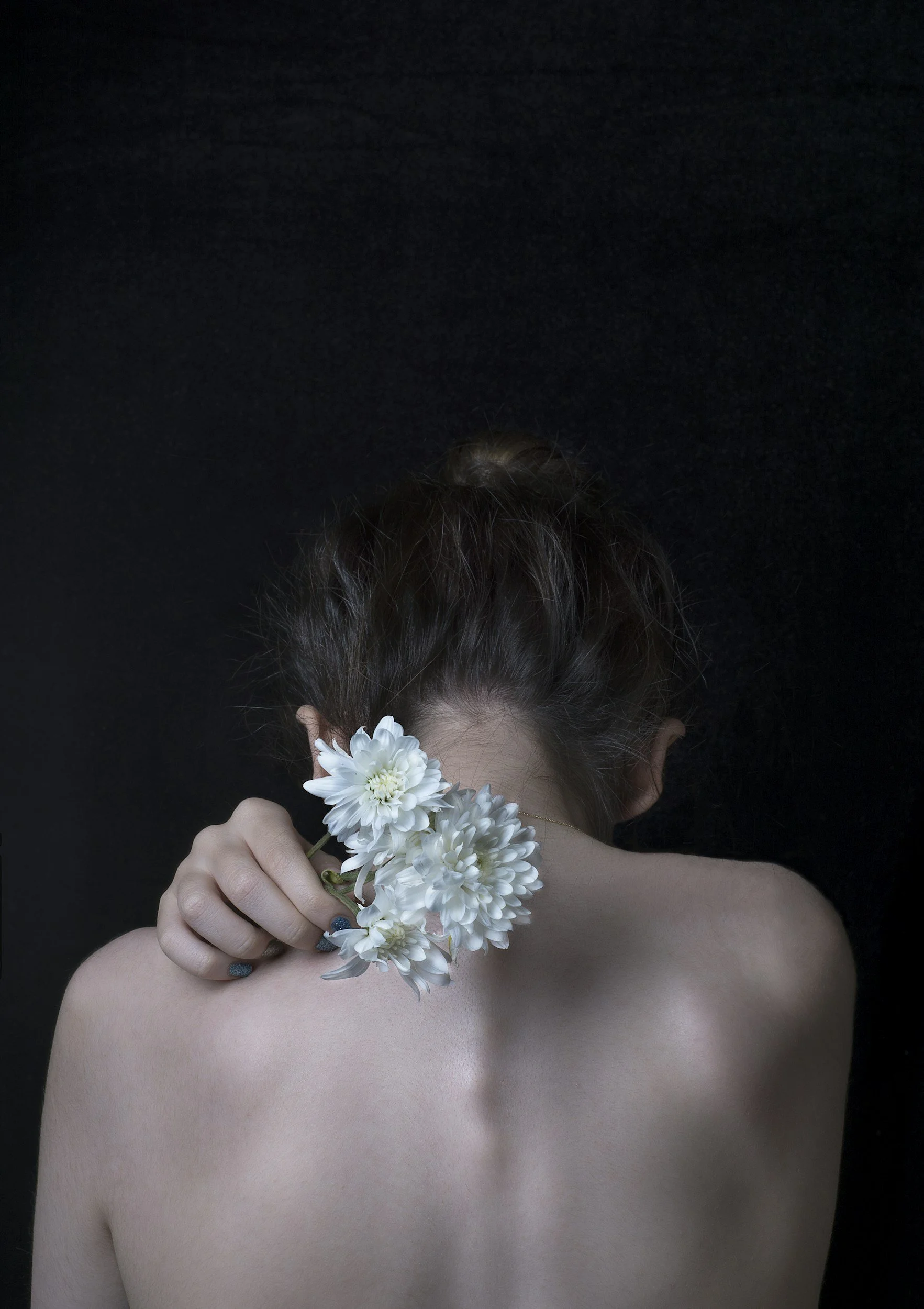 A woman with brown hair in a bun holding white flowers behind her head, facing away from the camera against a dark background