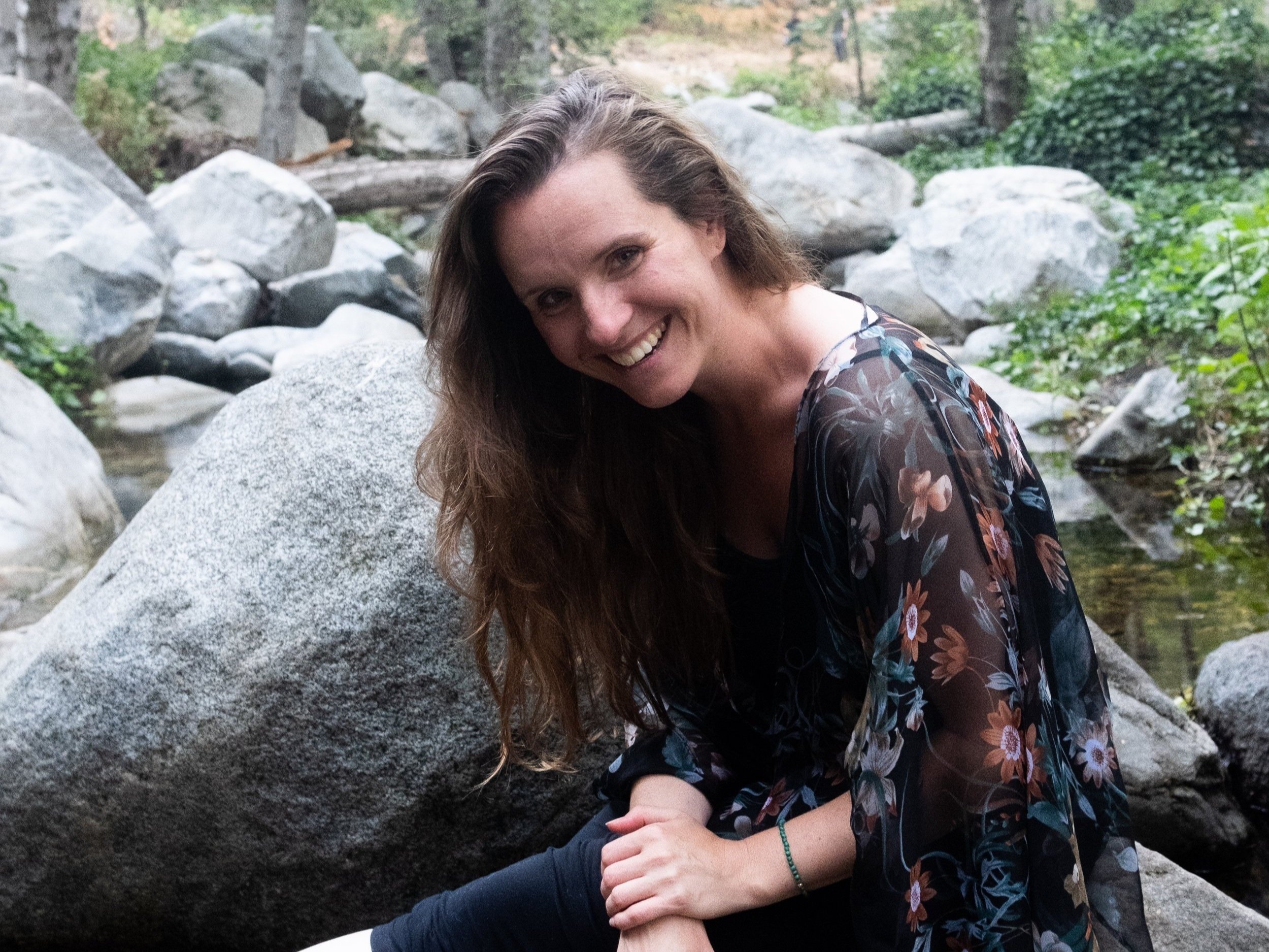 A woman with long brown hair smiling, sitting on a large rock near a creek in a forest setting with trees and greenery.