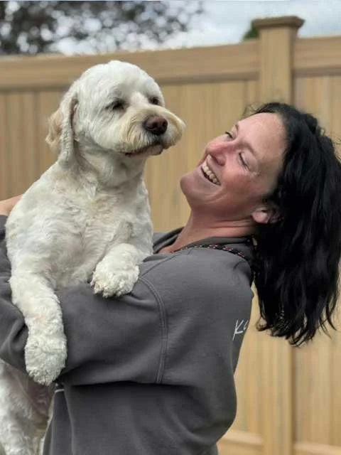 A woman with dark hair, holding up a white medium sized terrier mix leaning on her shoulder and smiling