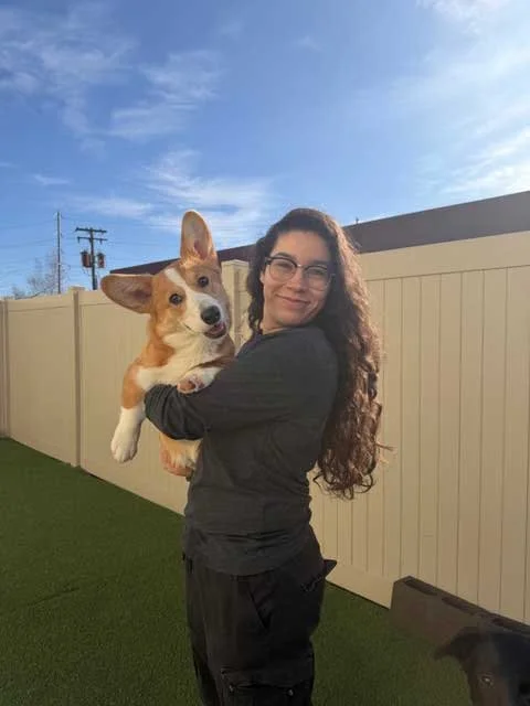 A woman with glasses and curly dark hair smiling next to a fluffy golden retriever dog with a happy expression in a fenced outdoor area.