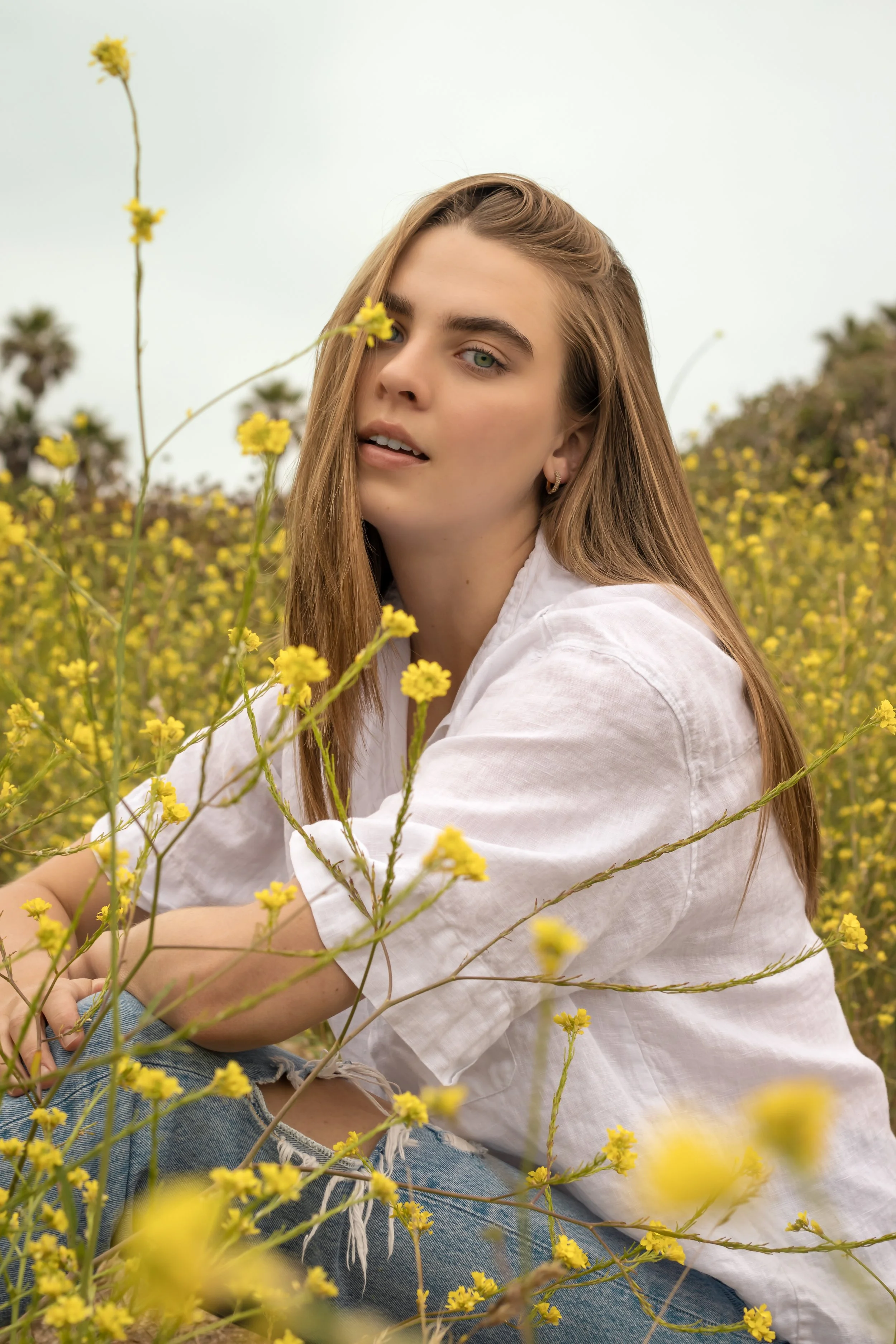 A young woman with long brown hair and green eyes sitting in a field of yellow flowers, wearing a white shirt and denim jeans, facing the camera with a soft expression.