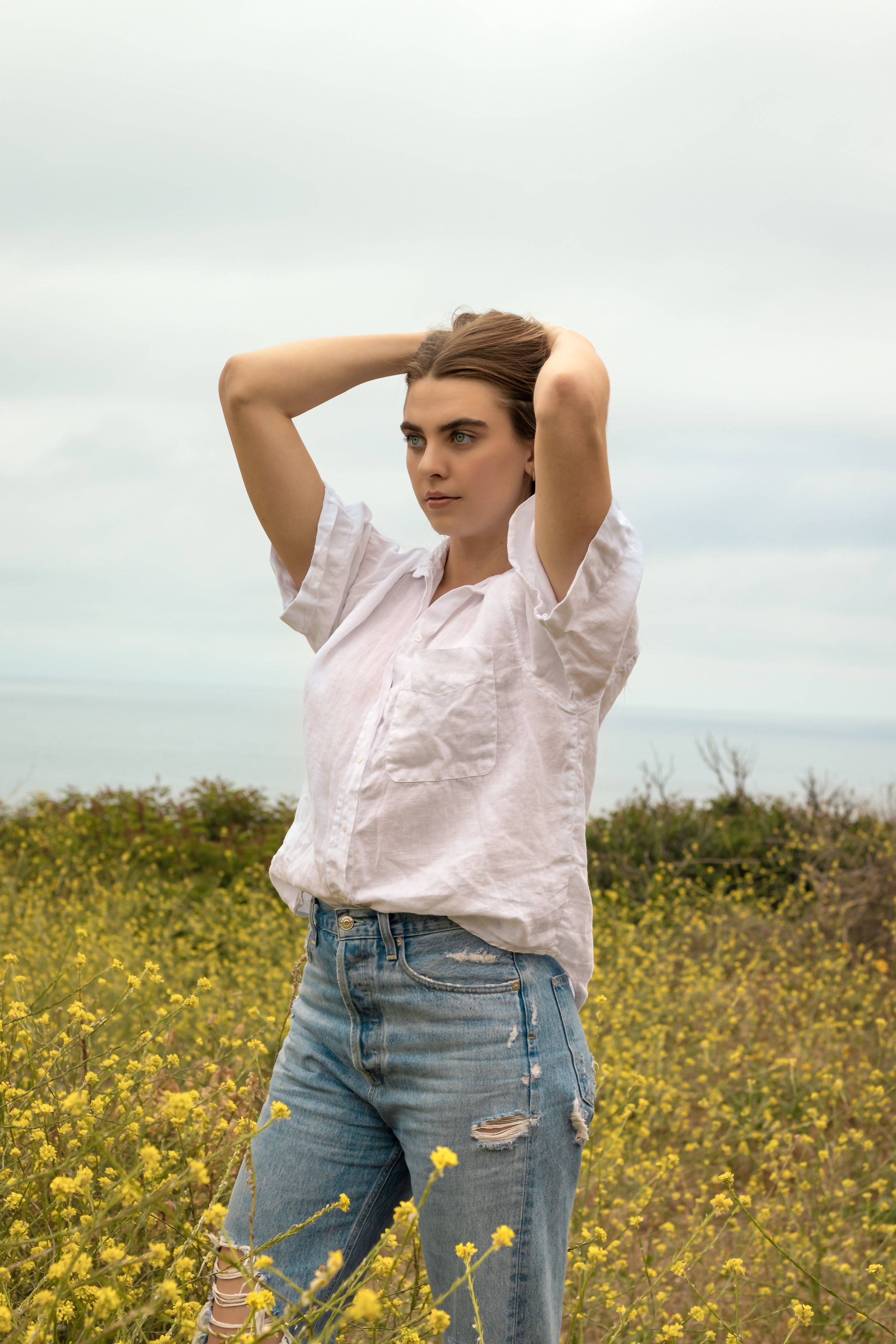 A woman standing in a field of yellow flowers with her hands behind her head, wearing a white shirt and ripped jeans, looking to the side with an overcast sky in the background.