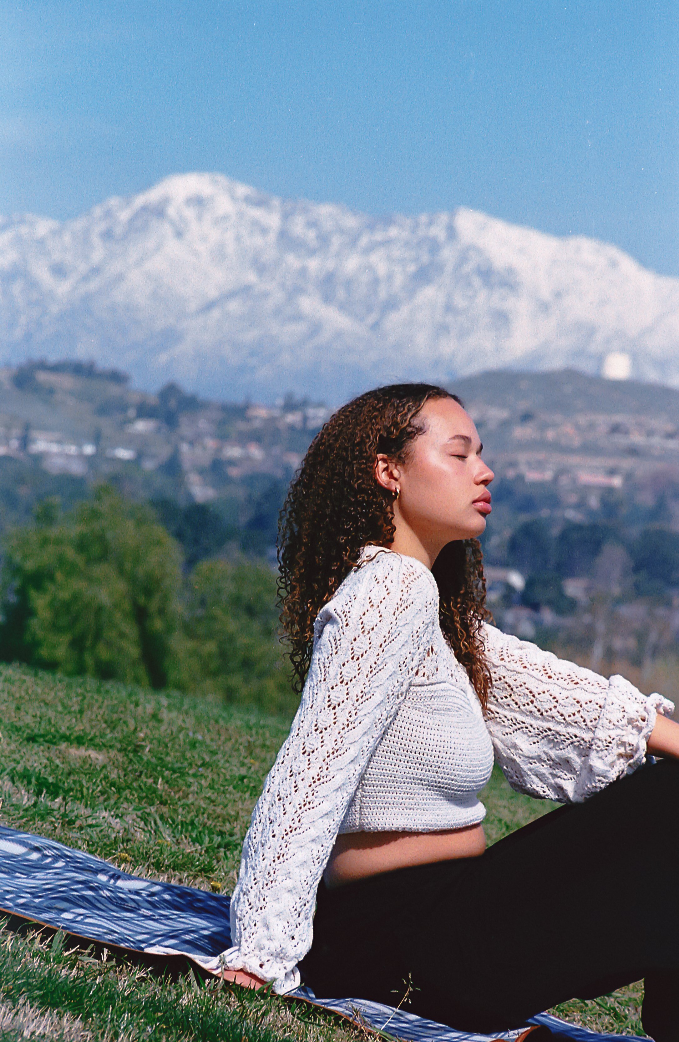 A young woman with curly hair sits on a grassy hill with her eyes closed, wearing a white knit top and black pants, with snow-capped mountains in the background.