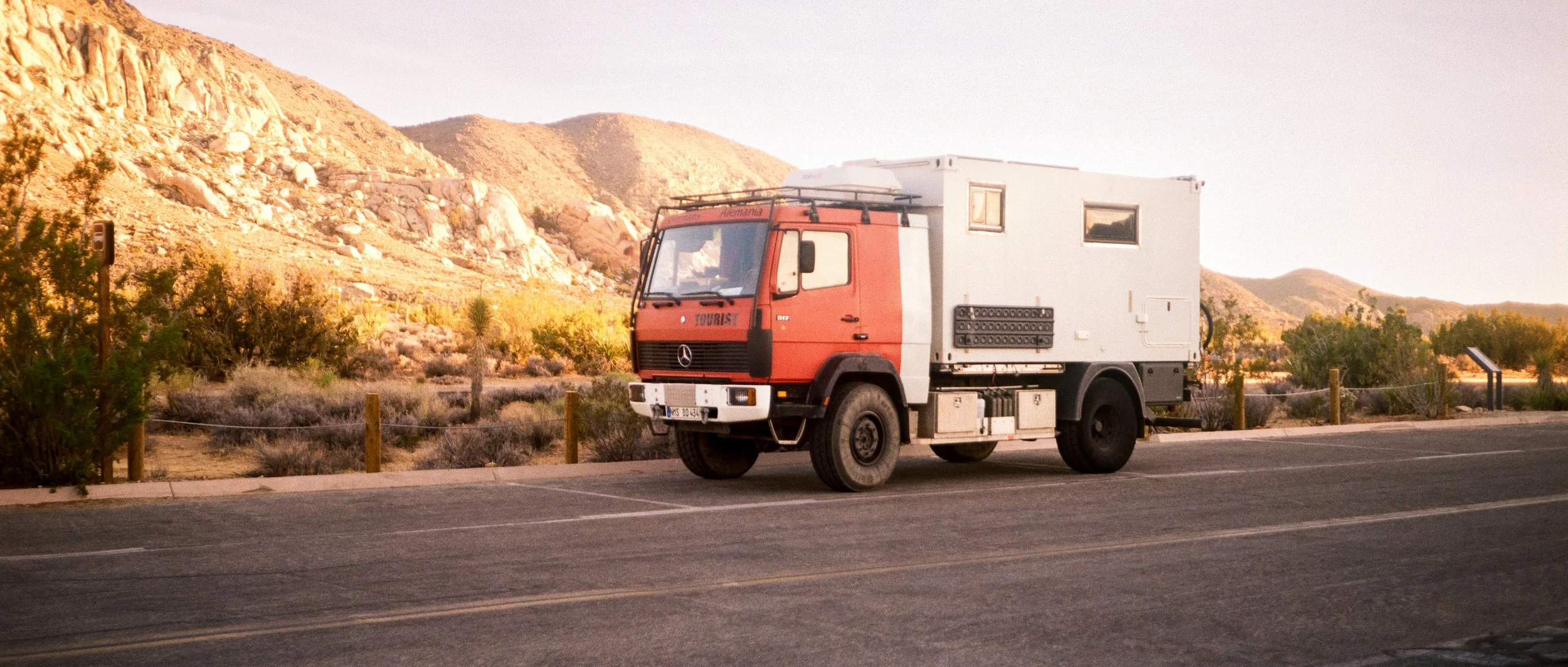 A vintage Mercedes-Benz utility truck parked along a desert road with rocky hills and sparse desert vegetation in the background.