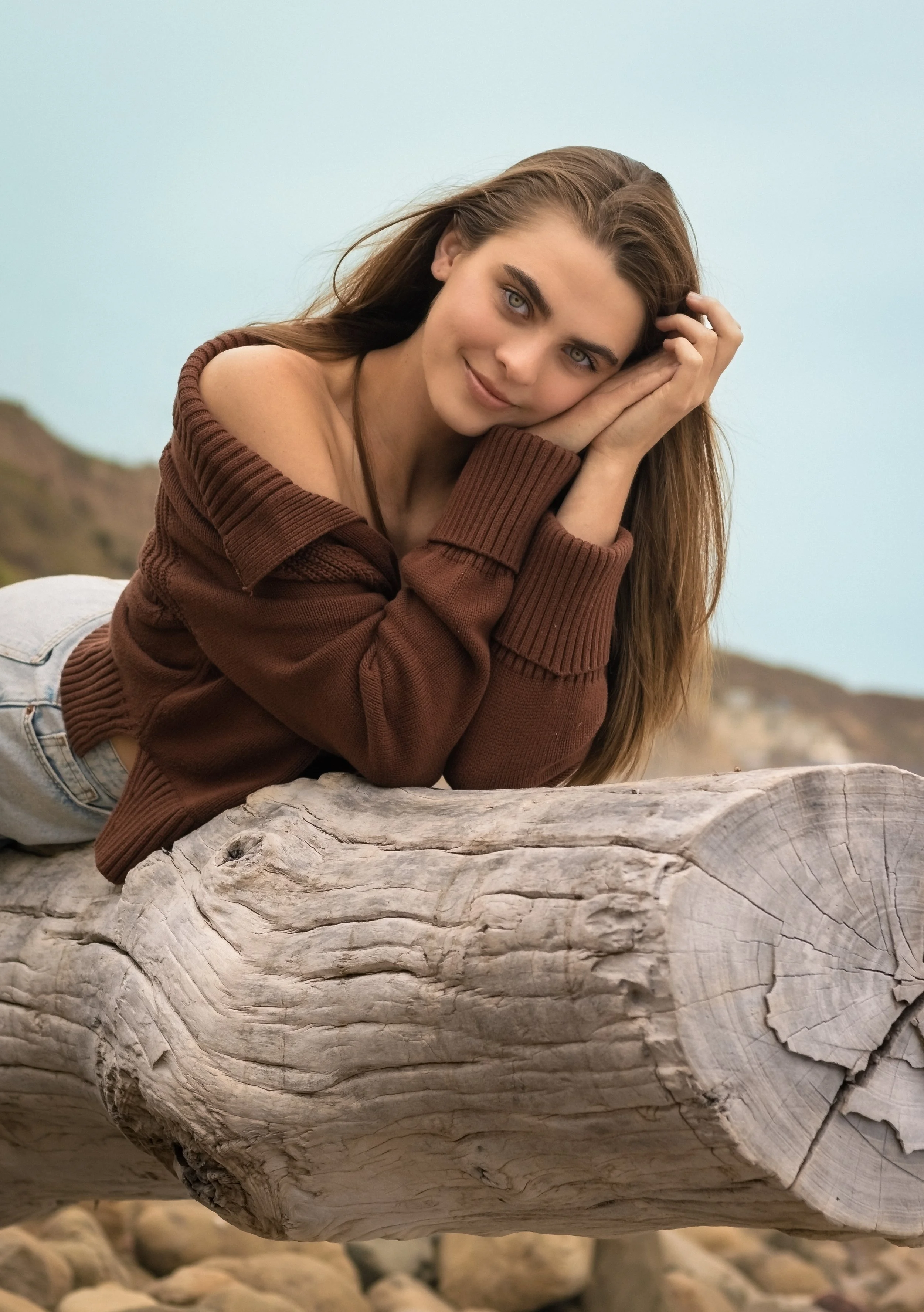 A young woman with long brown hair and blue eyes is lying on a large piece of driftwood outdoors, resting her head on her hands, and smiling gently at the camera. She is wearing a brown off-the-shoulder sweater and light jeans, with a background of a