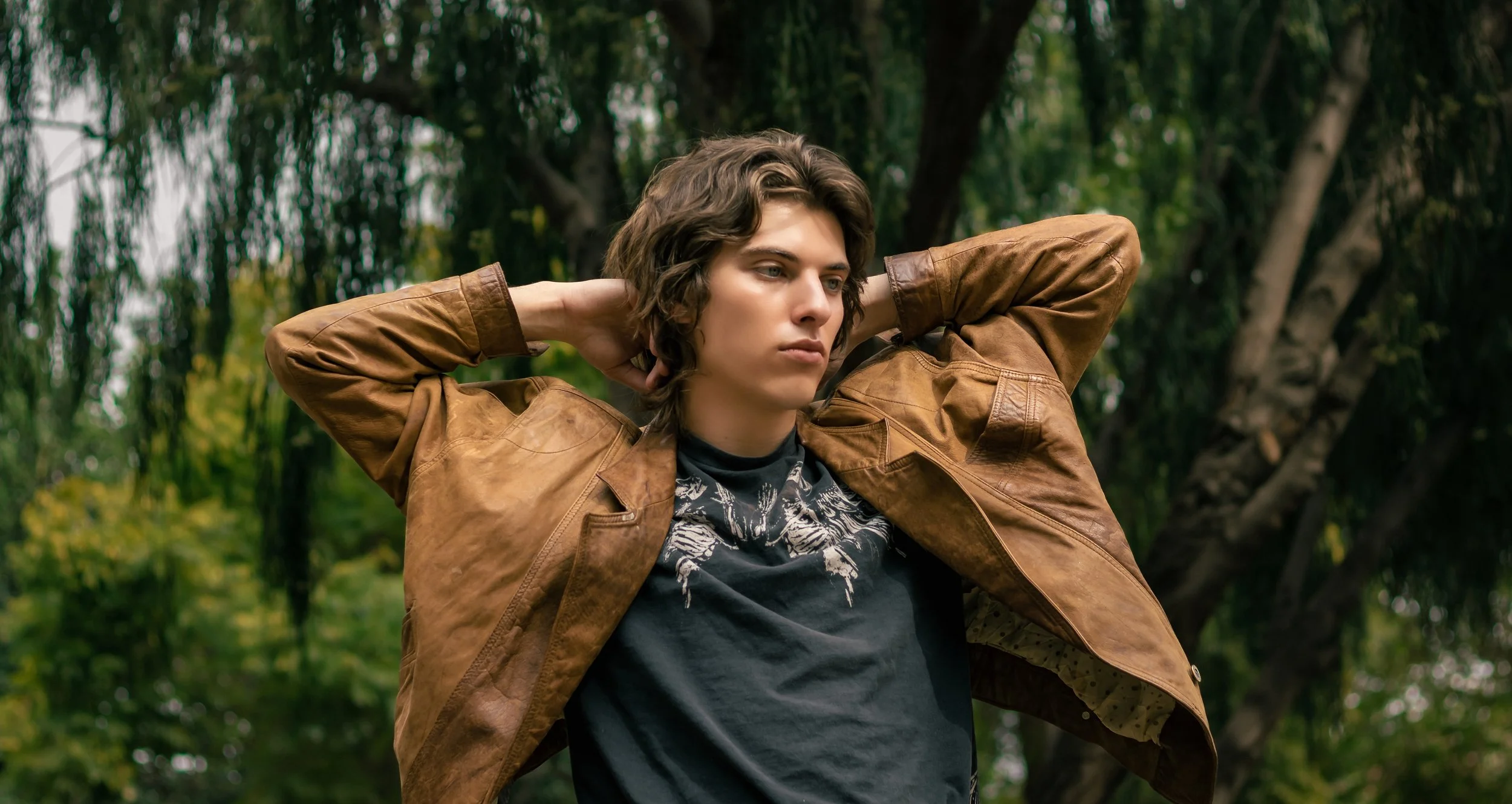A young person with shoulder-length wavy hair standing outdoors with trees in the background, wearing a brown leather jacket over a black graphic t-shirt, with their hands behind their head.