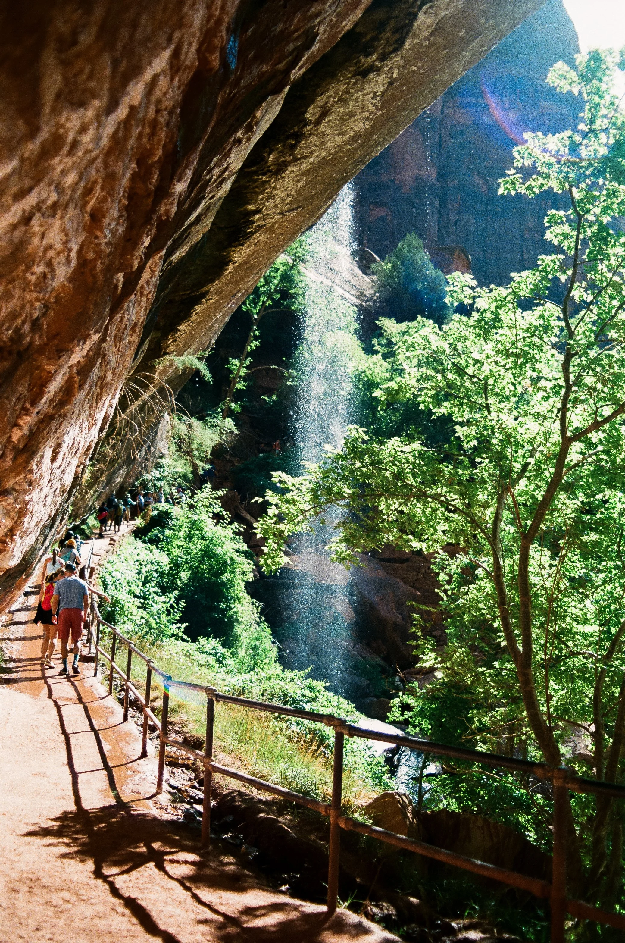 People walking on a trail next to a cliff with a vertical drop, overlooking a lush green forest and a waterfall in the background, with sunlight filtering through the trees.