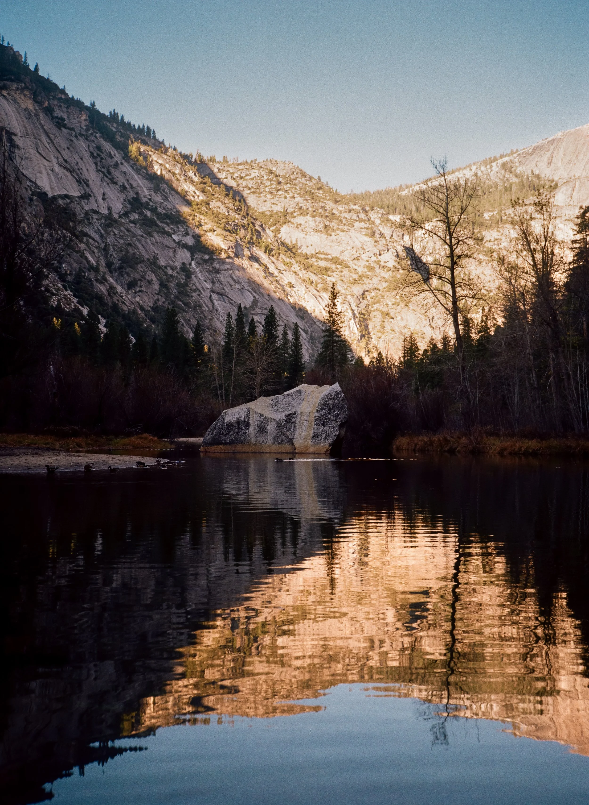 A serene mountain landscape with a calm river reflecting the trees and mountains, including a large boulder on the riverbank under clear skies.