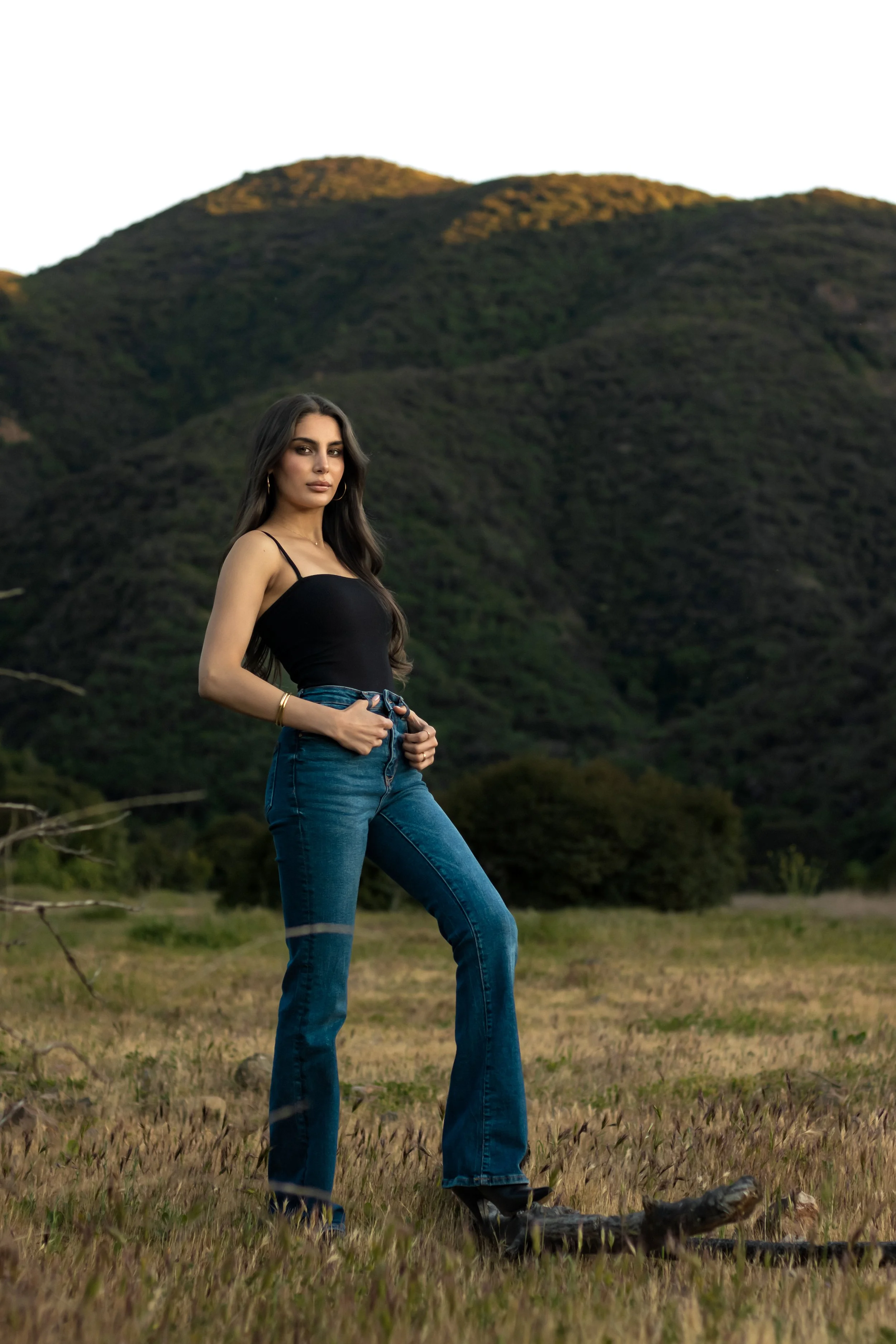 A woman stands in a grassy field with mountains in the background, wearing a black tank top and blue jeans.