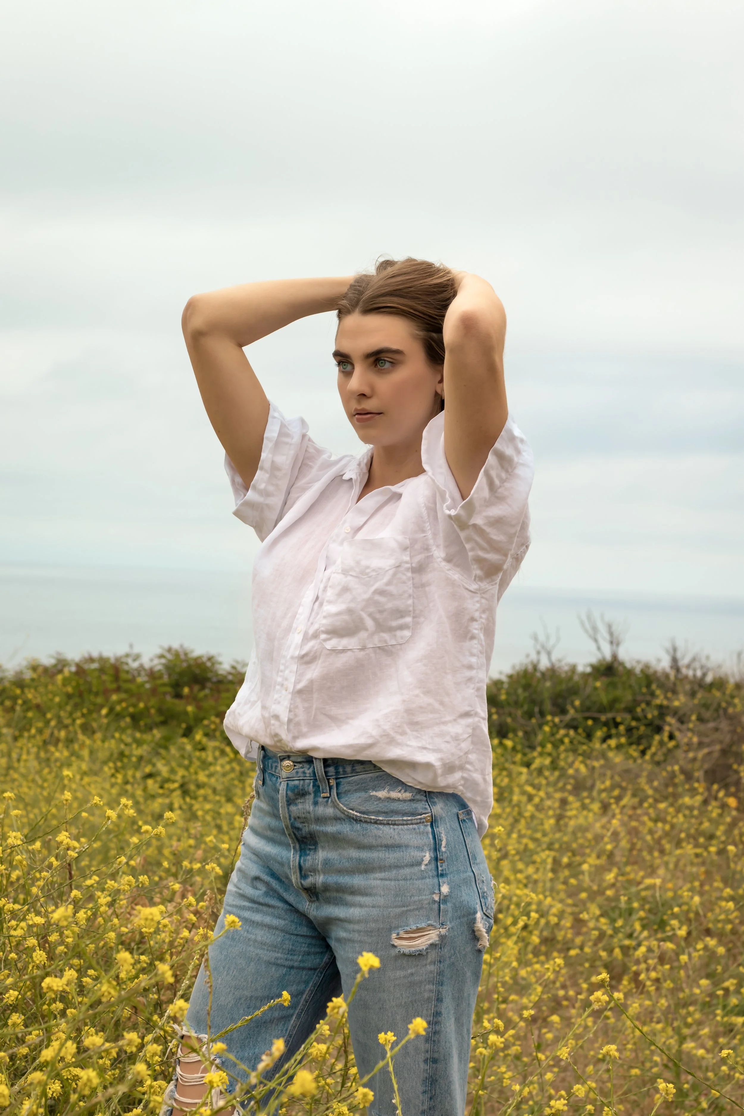 A young woman standing in a yellow flower field outdoors, wearing a white shirt and ripped jeans with her hands behind her head, looking into the distance.