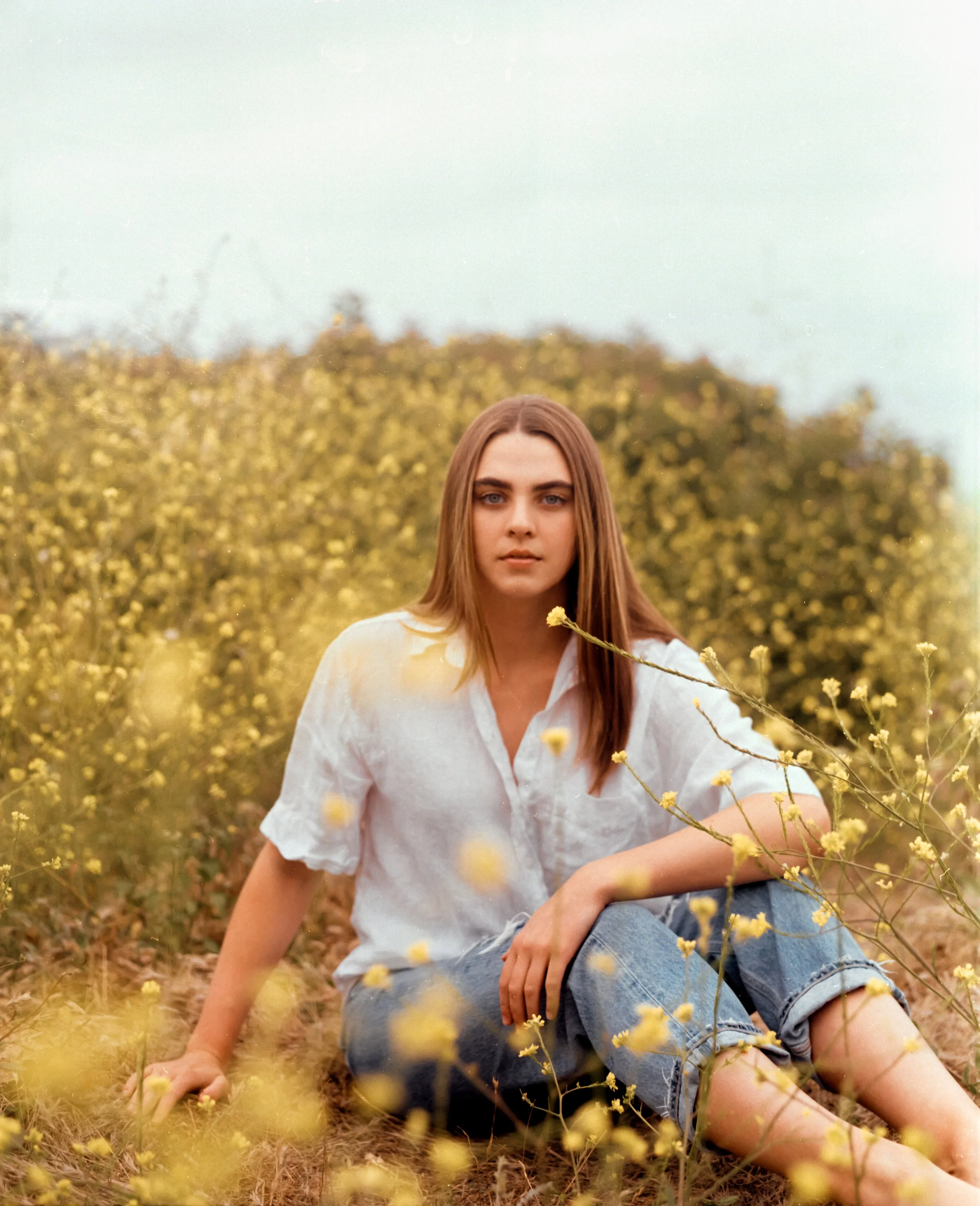 A young woman with long, straight brown hair sitting in a yellow flower field, wearing a white blouse and rolled-up jeans.