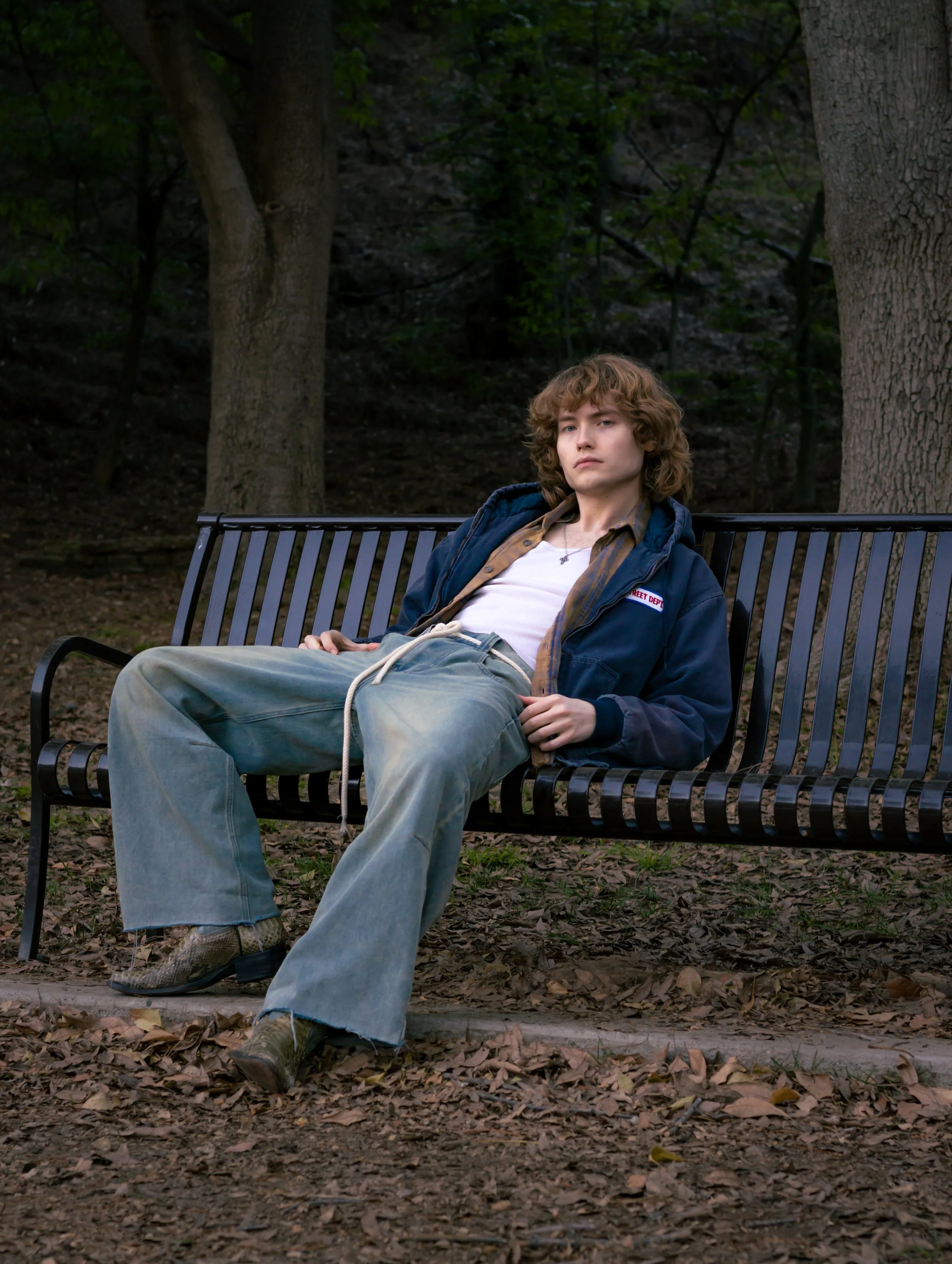 A young person with curly hair sitting on a park bench in a wooded area, wearing a blue jacket, beige shirt, and baggy jeans.