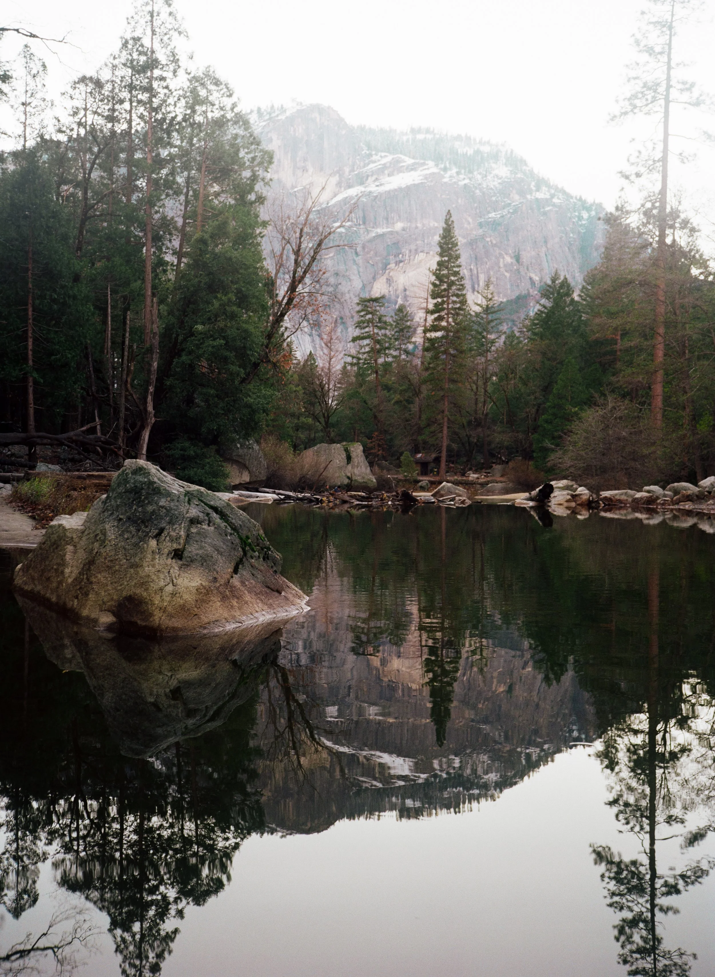 A tranquil lake surrounded by tall trees with a mountain in the background, and large rocks along the shoreline, reflected in the still water.