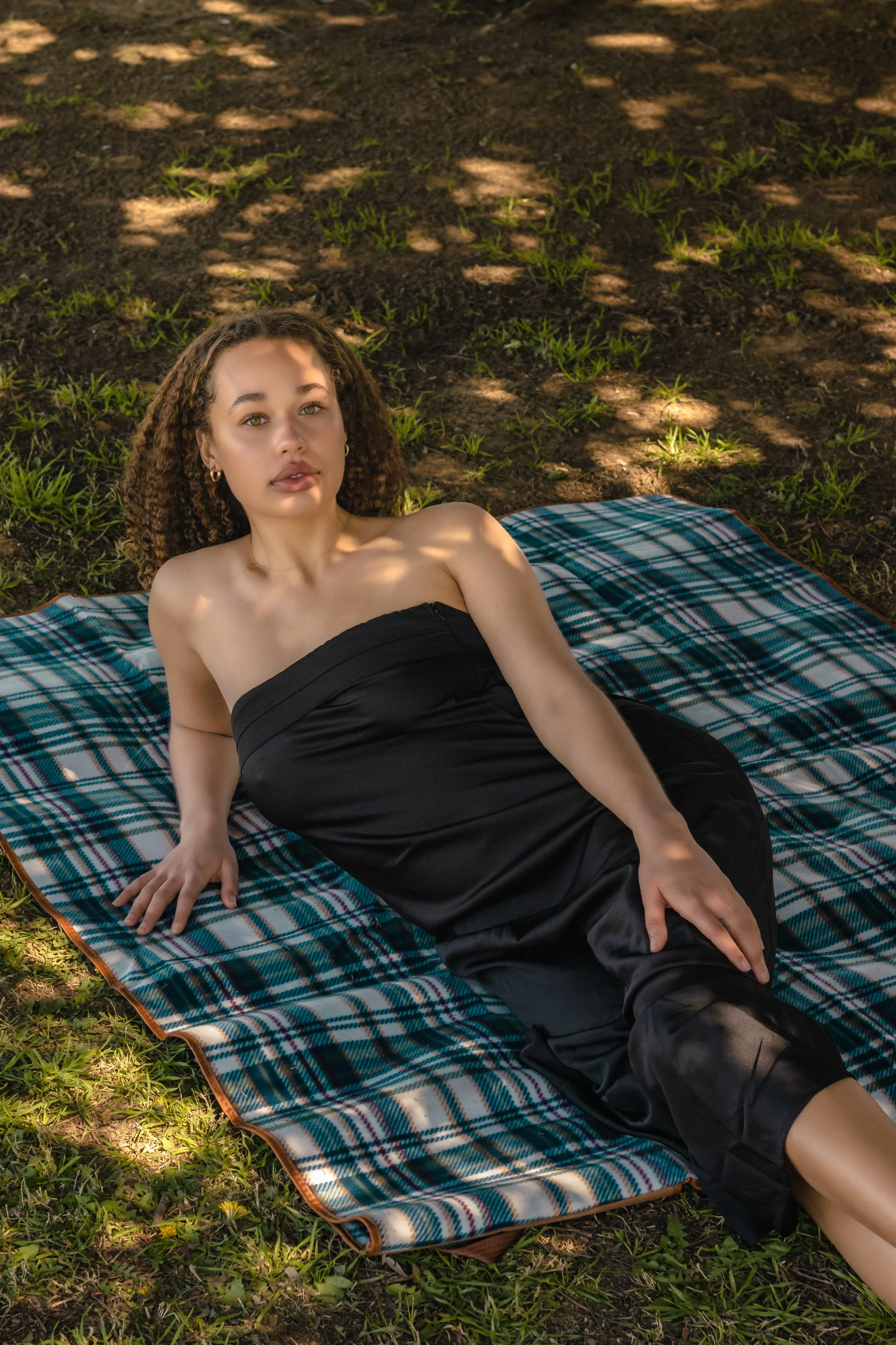 A young woman lies on a plaid blanket on the grass under shade trees, wearing a black strapless dress, looking at the camera.