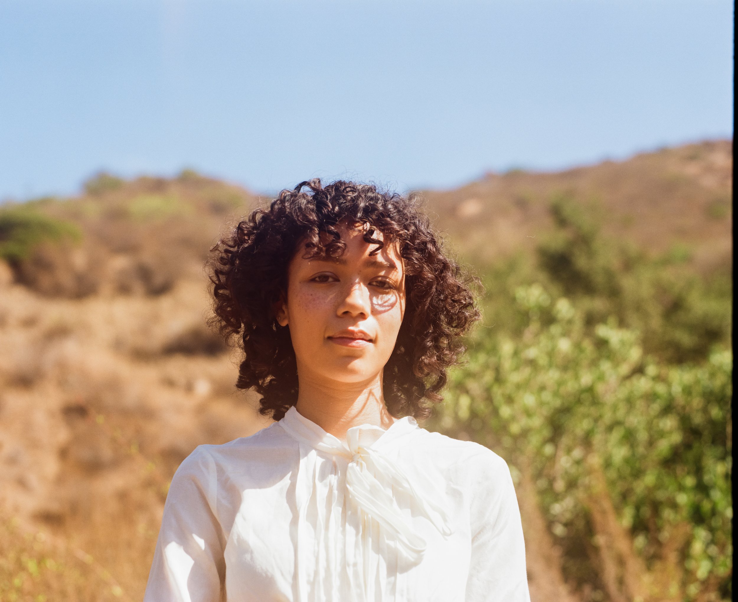 A woman with curly brown hair and light skin standing outdoors on a sunny day, wearing a white blouse with a bow tie, with a blurred background of trees and hills.