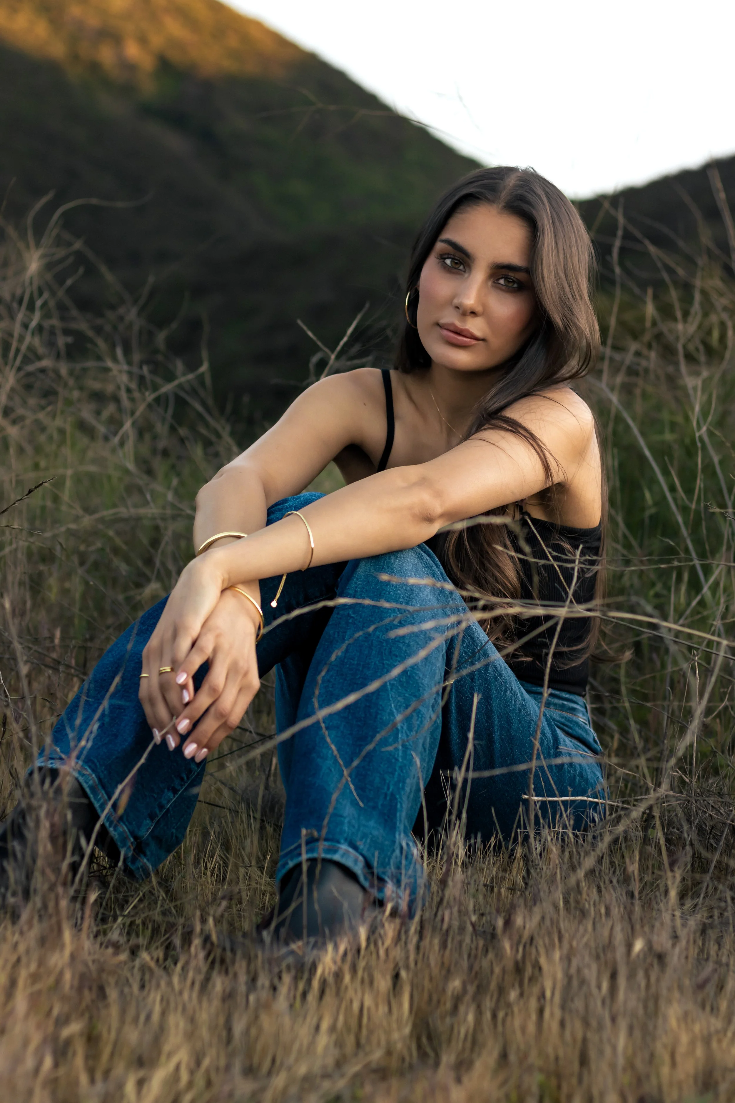 A young woman sitting in a grassy outdoor area during daytime, wearing a black tank top and jeans, with long dark hair and earrings, looking at the camera with a relaxed expression.
