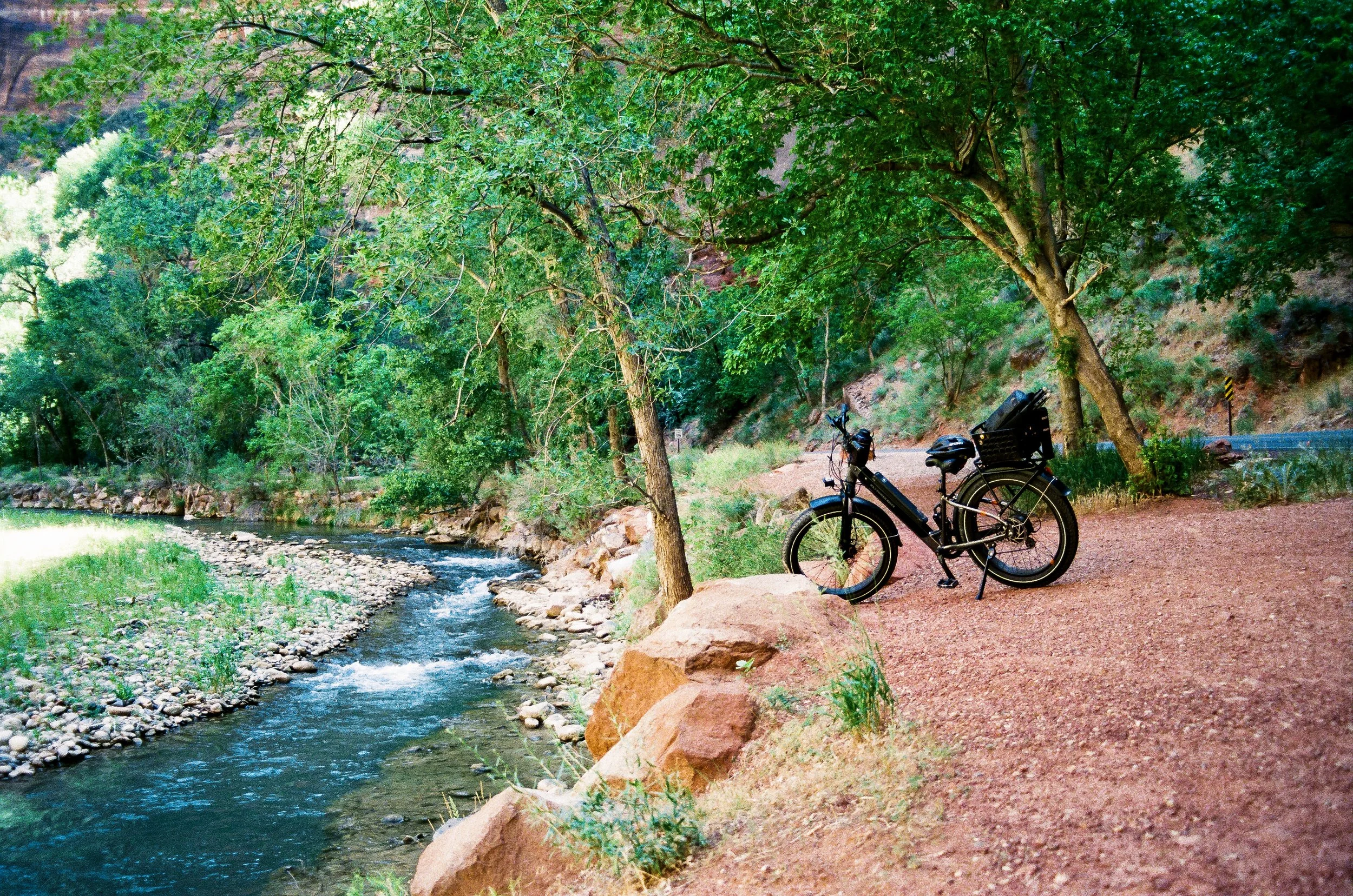 A black e-bike parked on a dirt path beside a small river, surrounded by green trees and rocky terrain.