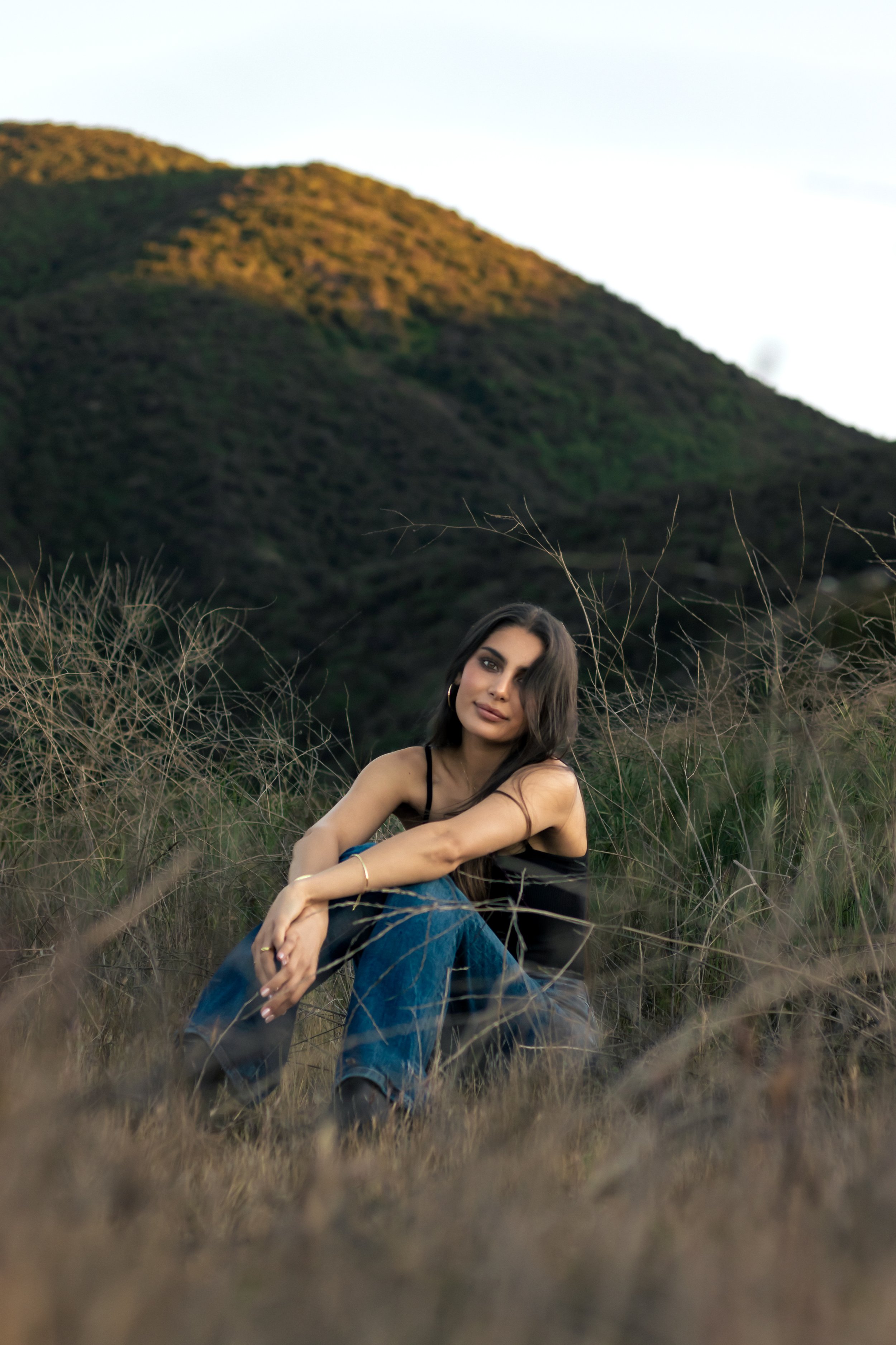 A woman with long dark hair sitting on dry grass in a field with hills in the background, wearing a black top and blue jeans.