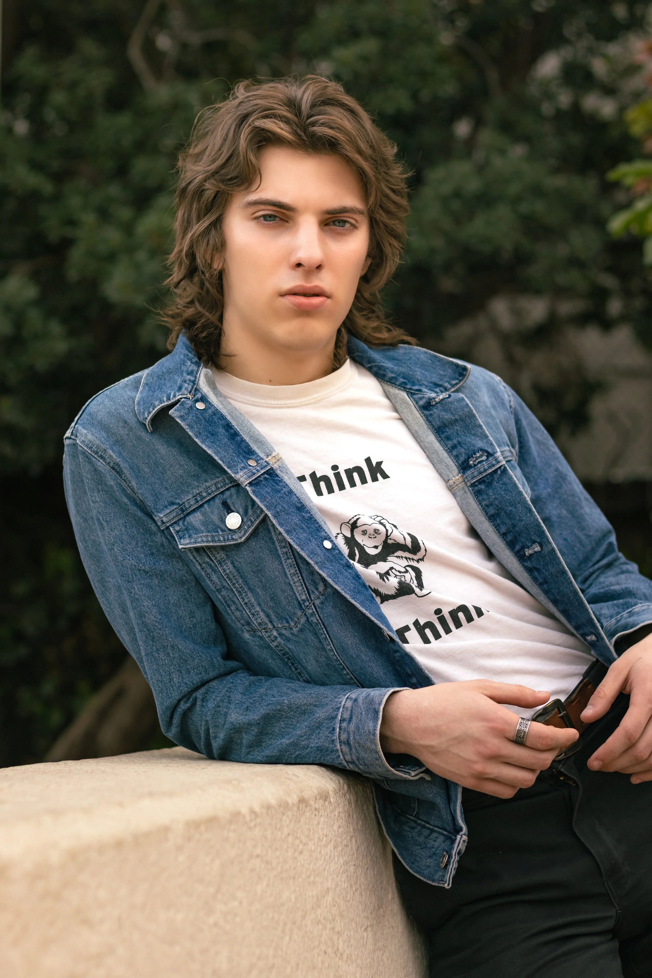 Young man with long, wavy brown hair wearing a denim jacket over a white graphic T-shirt with a monkey and the words 'Think Think.' standing outdoors with greenery in the background.