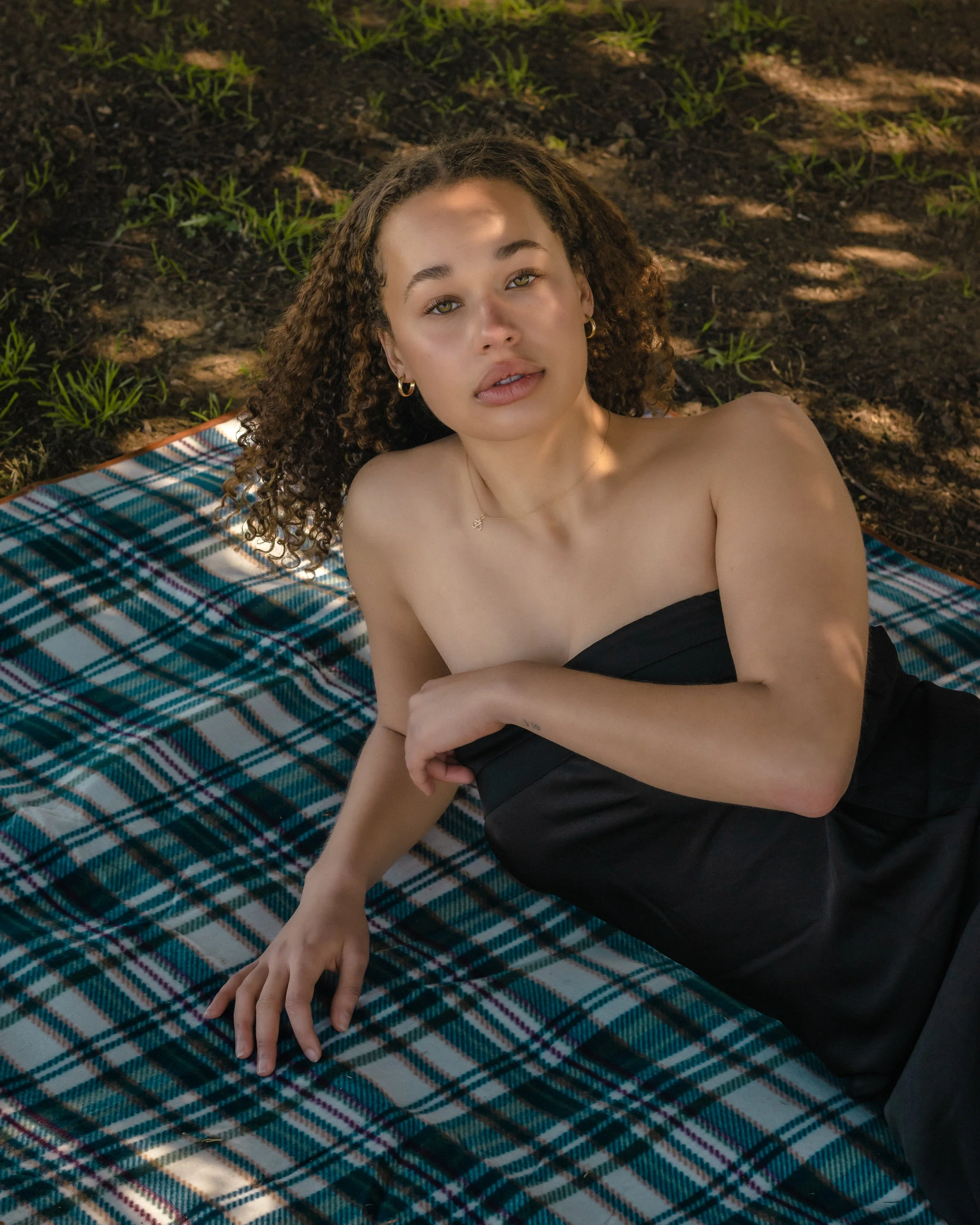 A young woman with curly hair lying on a checkered blanket outdoors, looking at the camera.