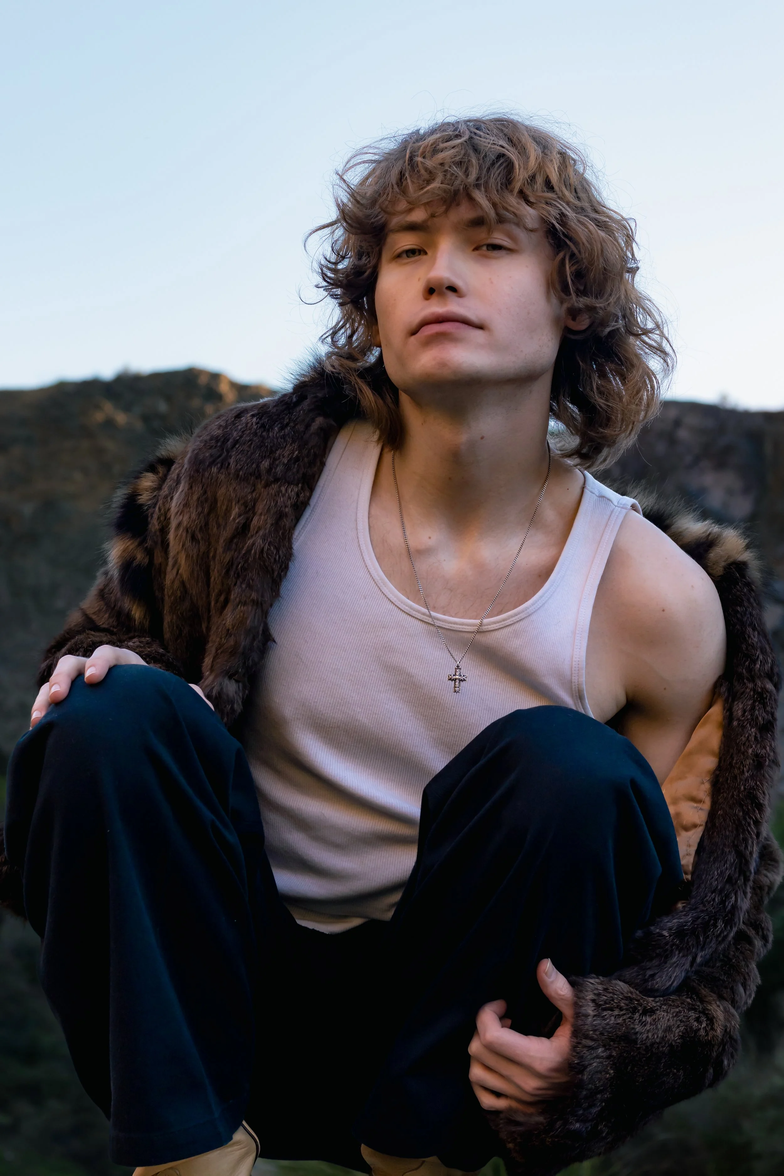 A young man with curly hair wearing a white tank top, a fur coat, and a necklace with a cross, crouching outdoors against a mountain landscape during dusk.