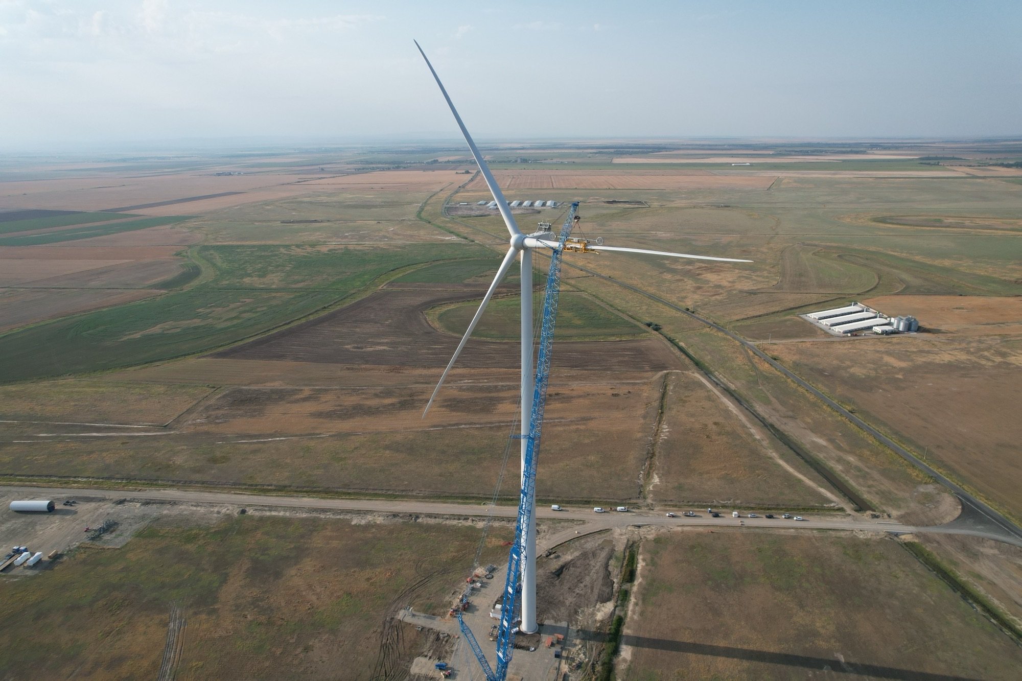 An aerial view of a large wind turbine under construction in a rural area, with a tall crane supporting the turbine and several vehicles nearby. The landscape consists of fields and farmland.