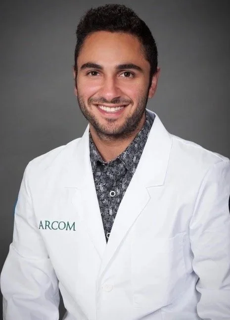 A male doctor with short dark hair, beard, and light skin, wearing a white medical coat with ARCOM embroidered on it, and a patterned shirt underneath, smiling against a gray background.