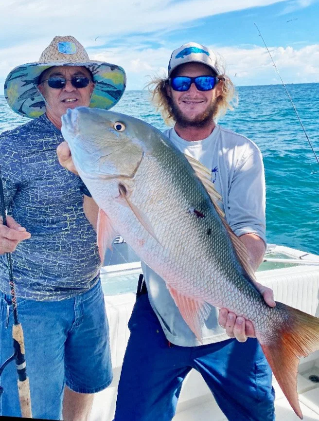 Two men on a boat, one holding a large fish, with the ocean and blue sky in the background.