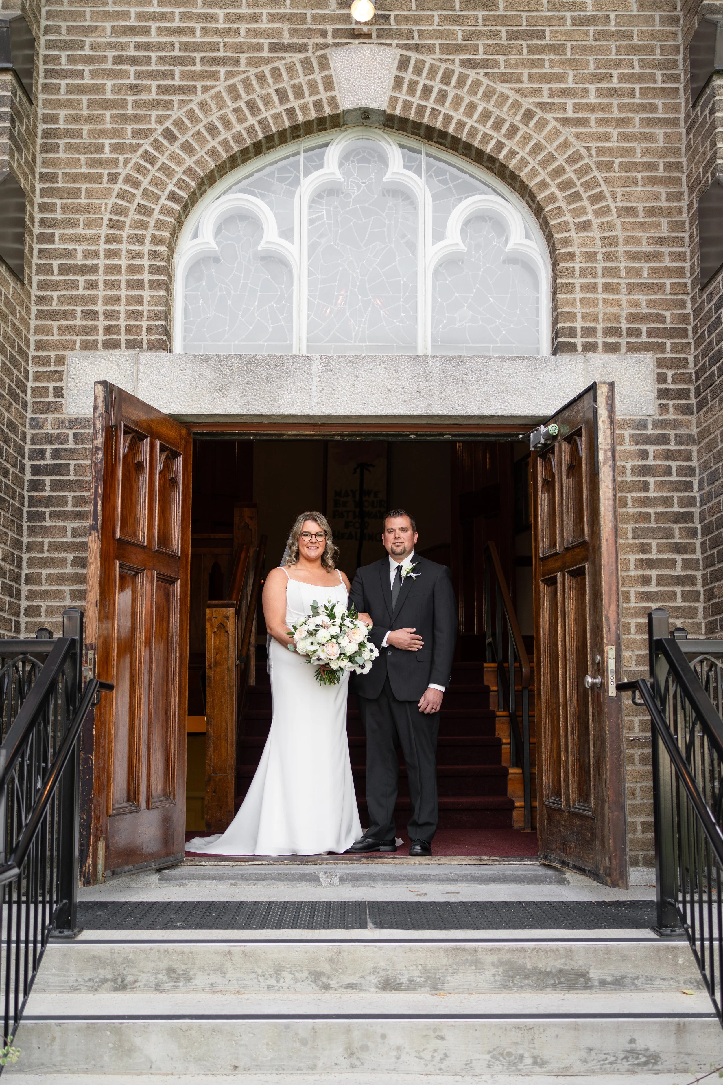 A bride and groom standing in the church entrance, the bride holding a bouquet of white roses and greenery, both smiling for a portrait. Pembroke church