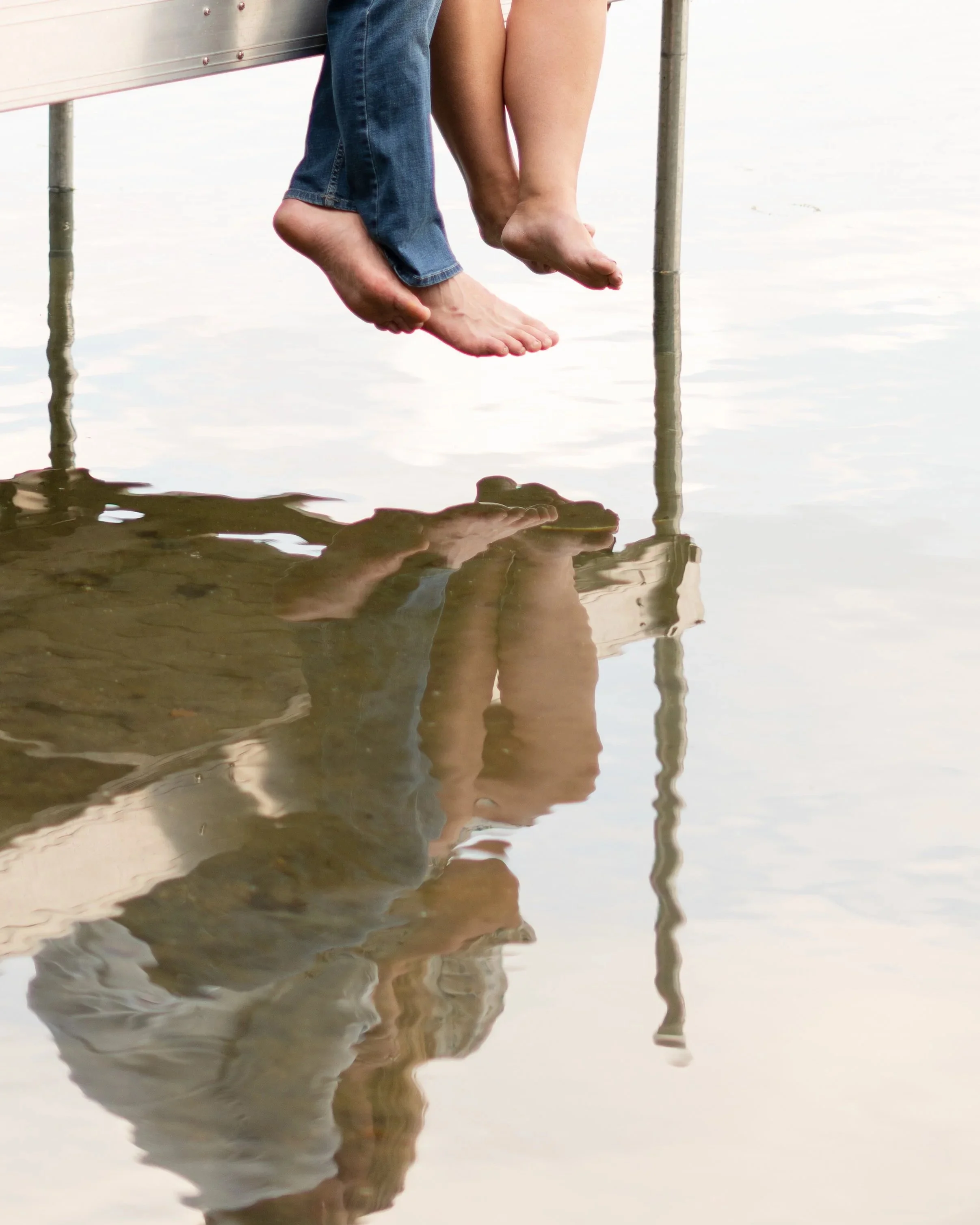 Close-up of a couple sitting on a dock with feet dangling above the water, reflecting in the water.