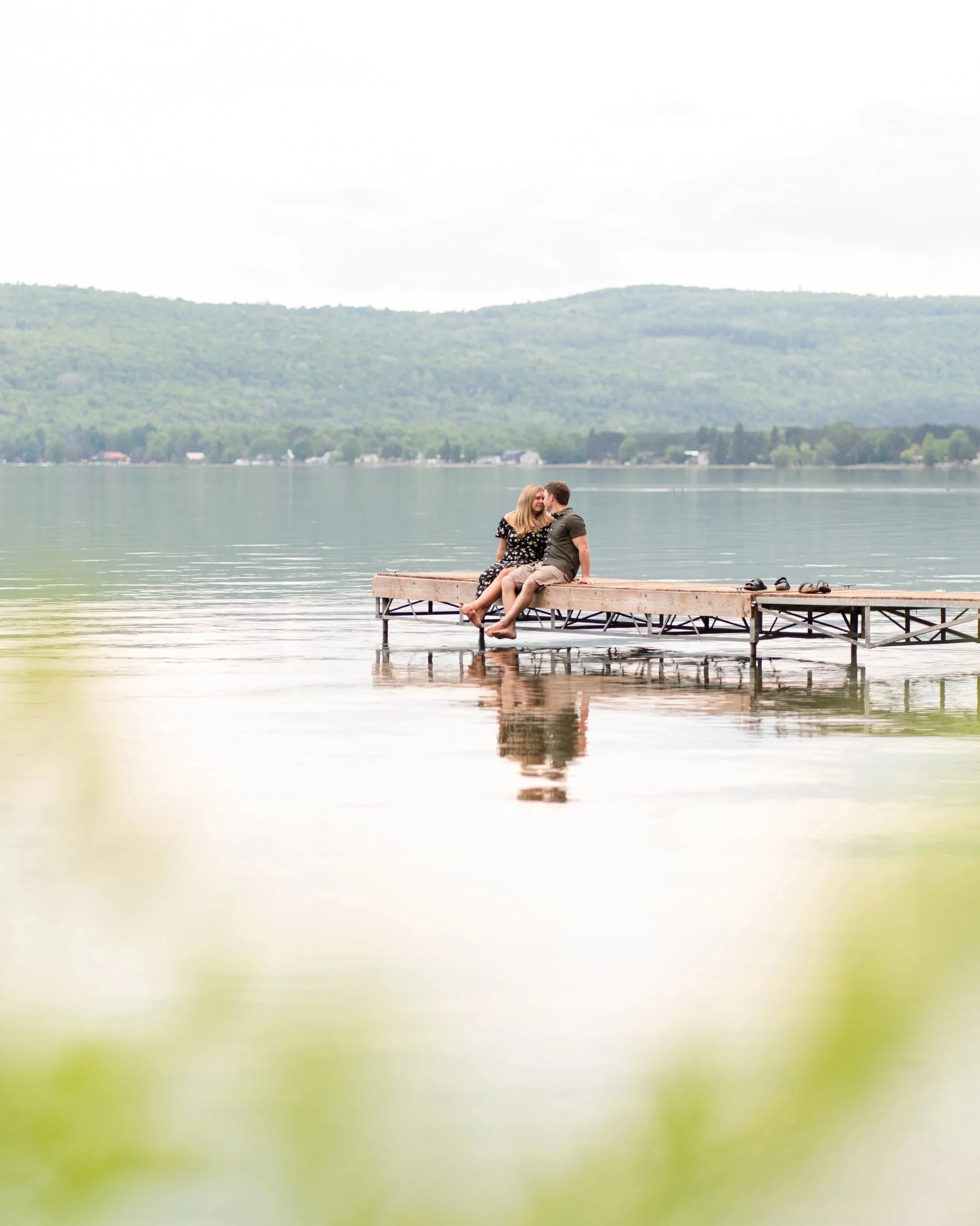 A couple sitting on a wooden dock by a lake, sharing a kiss, with mountains in the background.