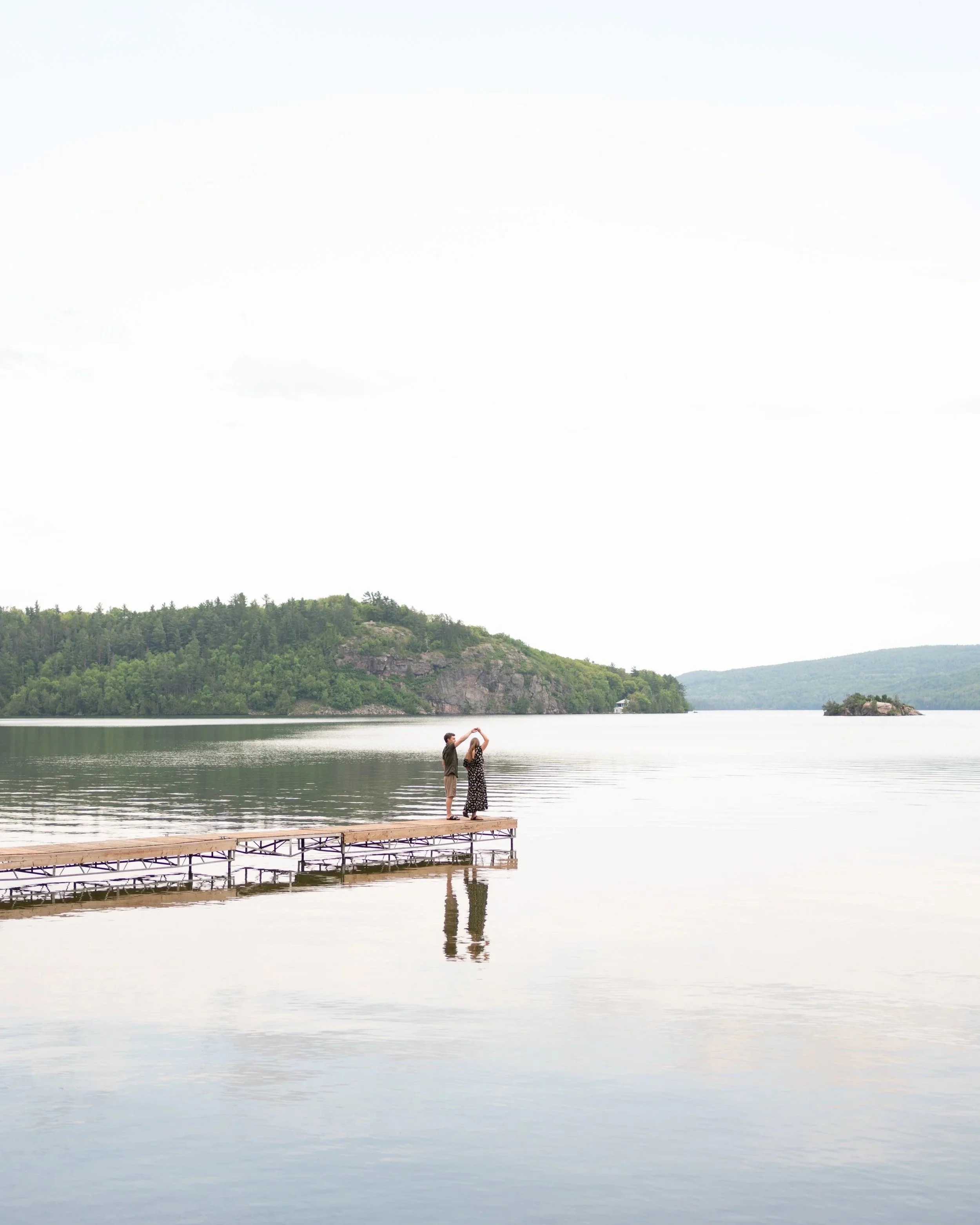 A couple stands on a wooden dock extending into calm water, holding hands as they look at each other with a scenic backdrop of green hills and a cloudy sky.