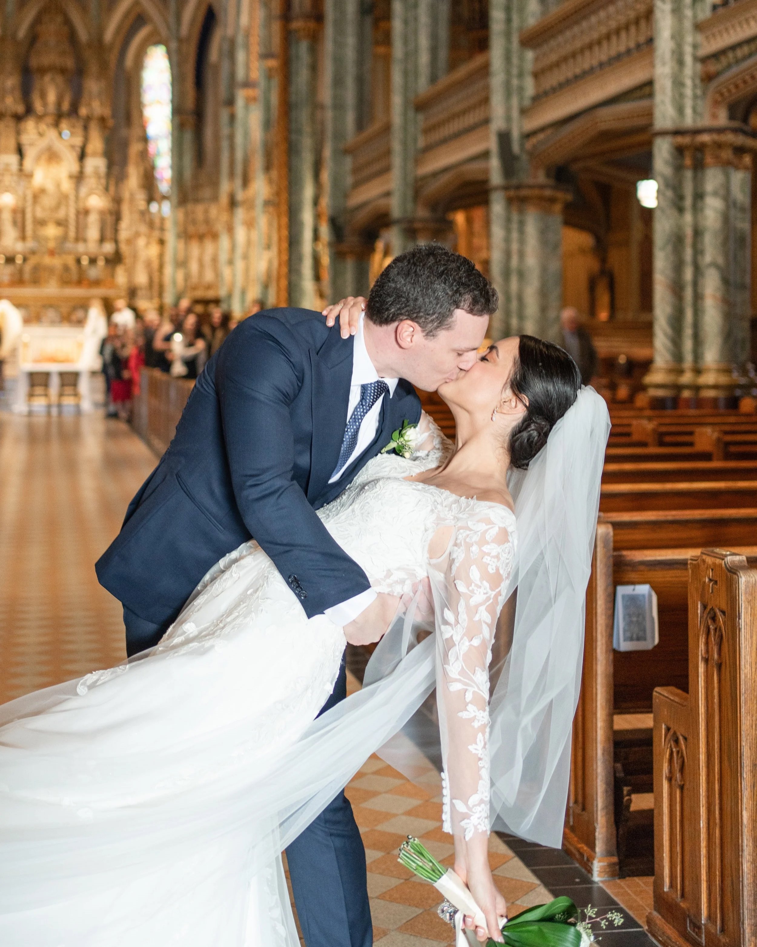 A newlywed couple sharing a kiss in a church, with the groom dipping the bride. The bride holds a bouquet and wears a lace wedding dress with a veil, while the groom is in a navy suit. The church interior features ornate columns and stained glass win