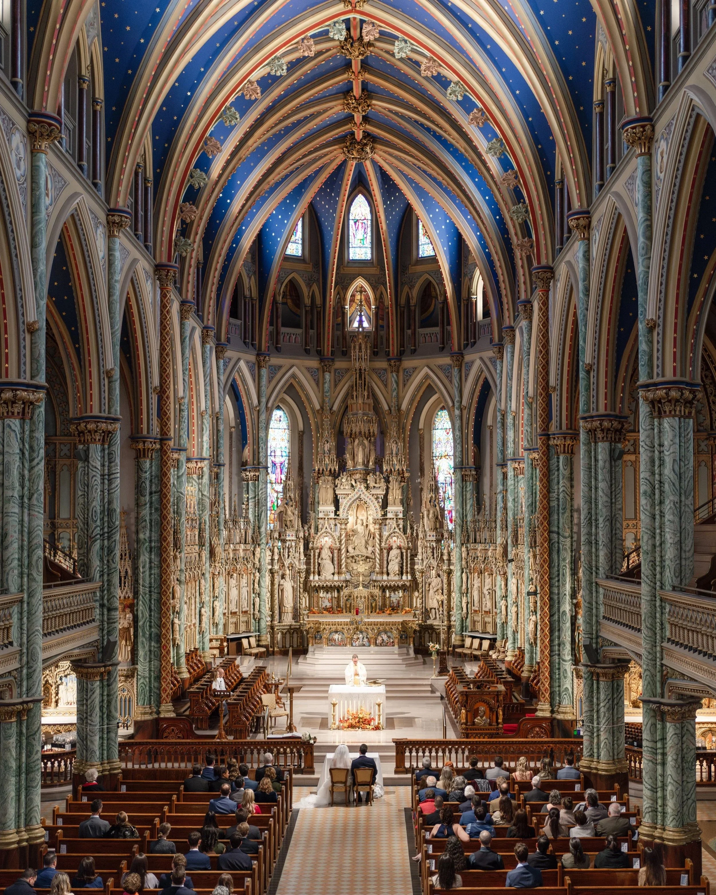 A wedding ceremony taking place inside a grand, ornate church with high vaulted ceilings, decorated with blue and gold accents, stained glass windows, intricate sculptures, and religious artwork. The bride and groom are seated at the altar, facing a 